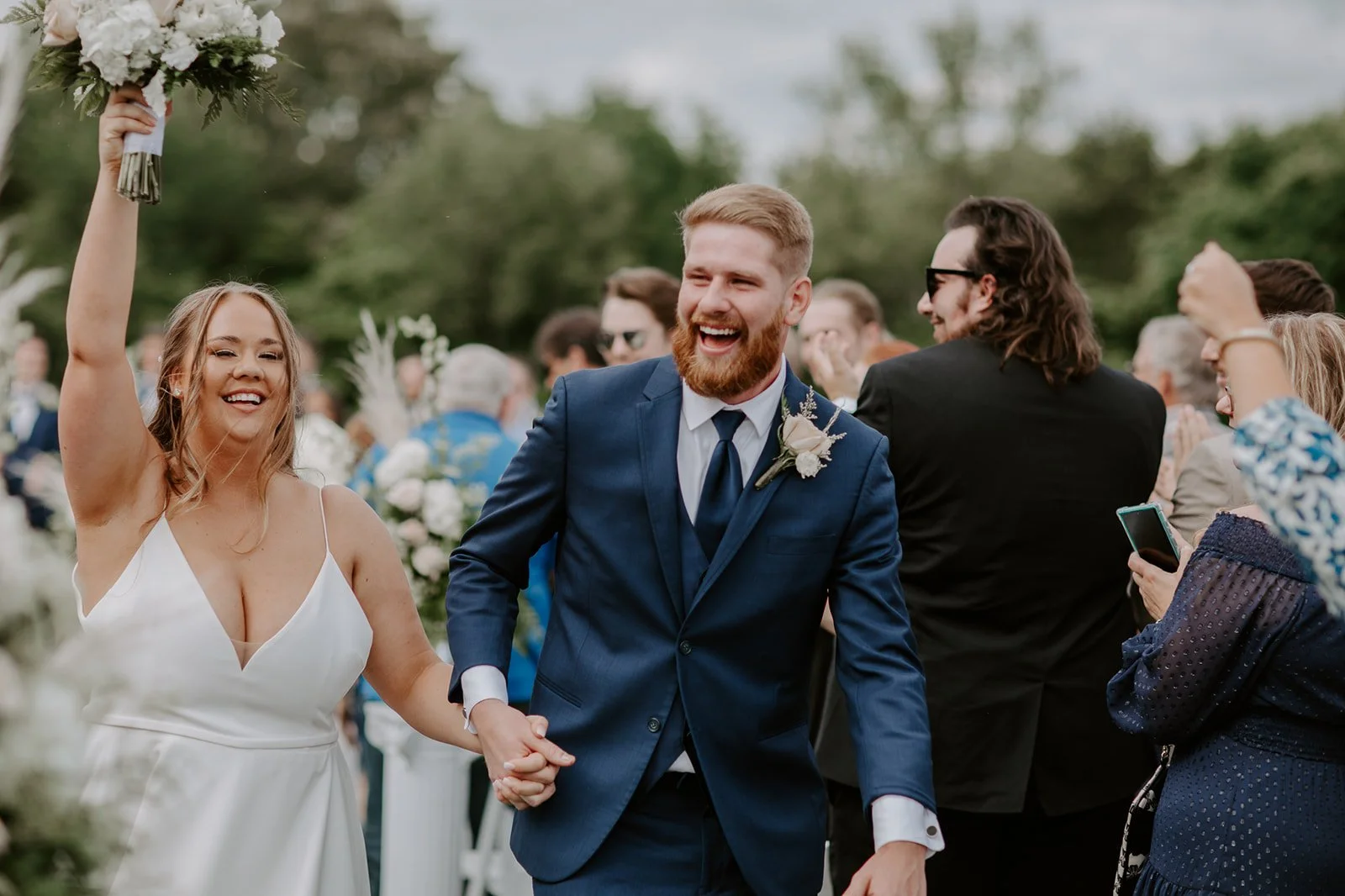 A joyful bride holding a bouquet and a groom in a blue suit walking hand in hand, celebrating outdoors at a wedding ceremony with smiling guests in the background.