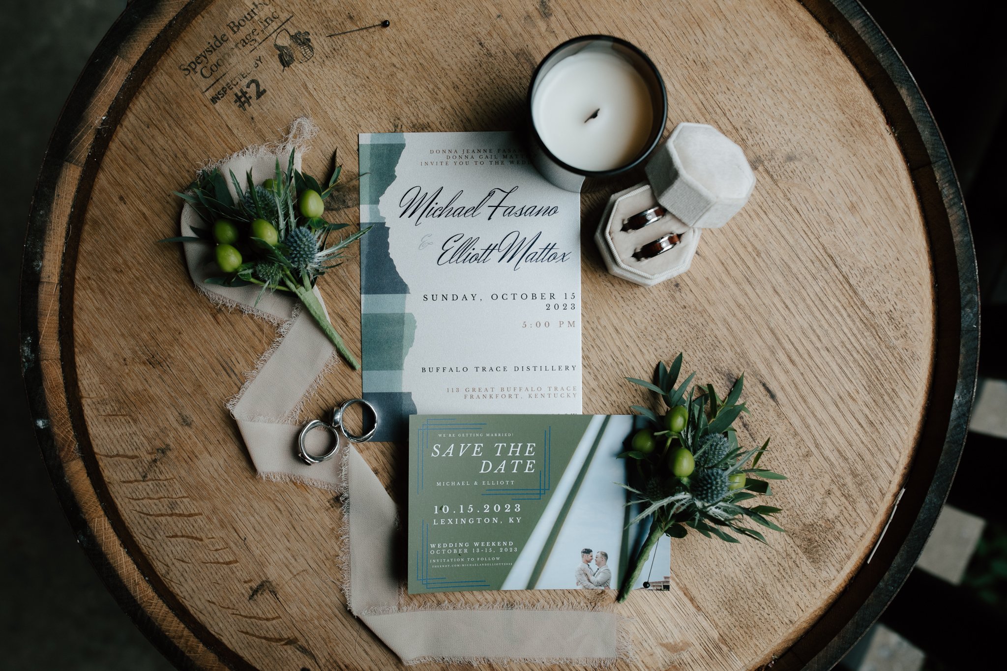 Wedding invitation and save-the-date card on a wooden barrel, with greenery, wedding rings in a box, and a candle.