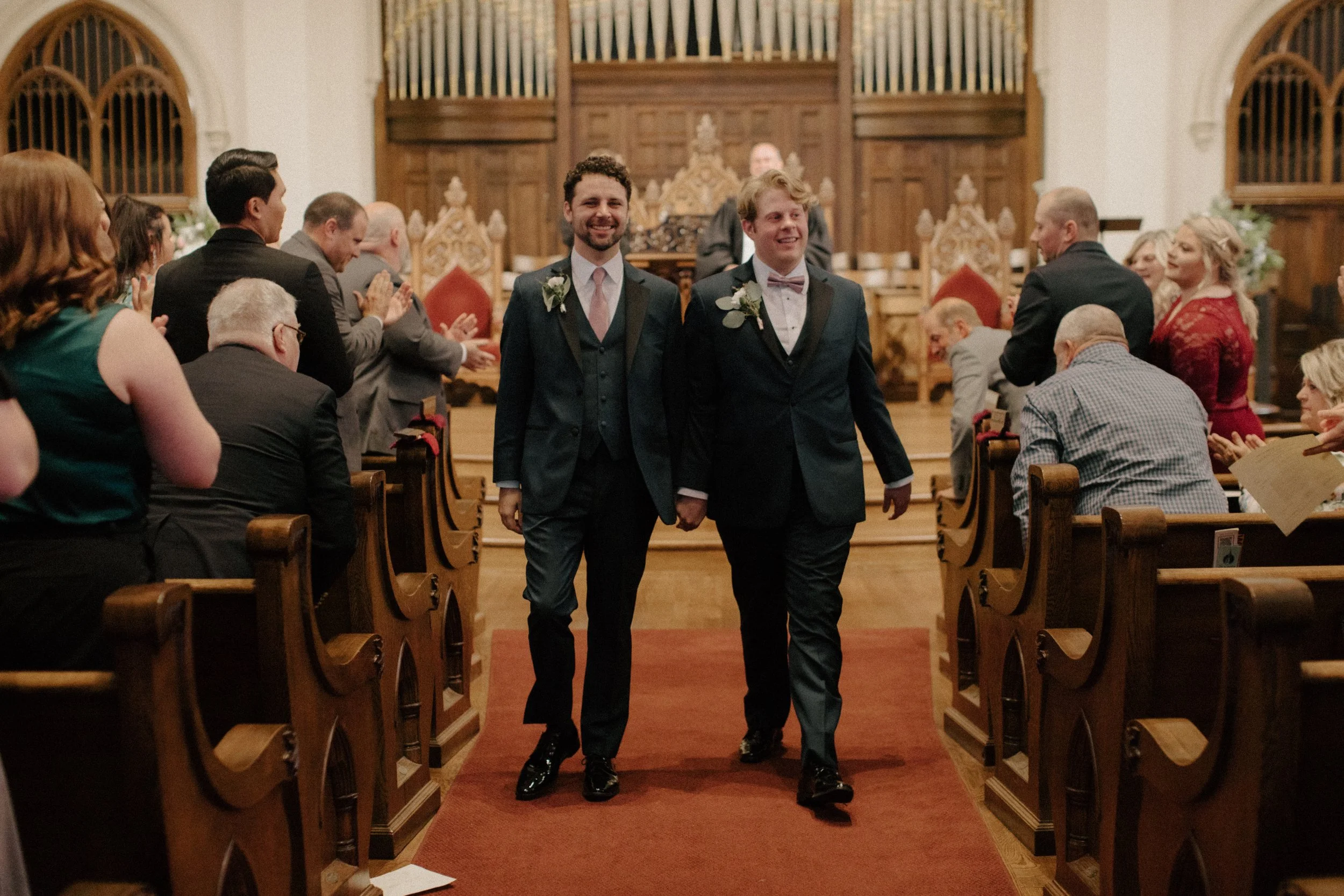 Two grooms walking down the aisle in a church after their wedding ceremony, holding hands and smiling, with guests applauding from the pews.