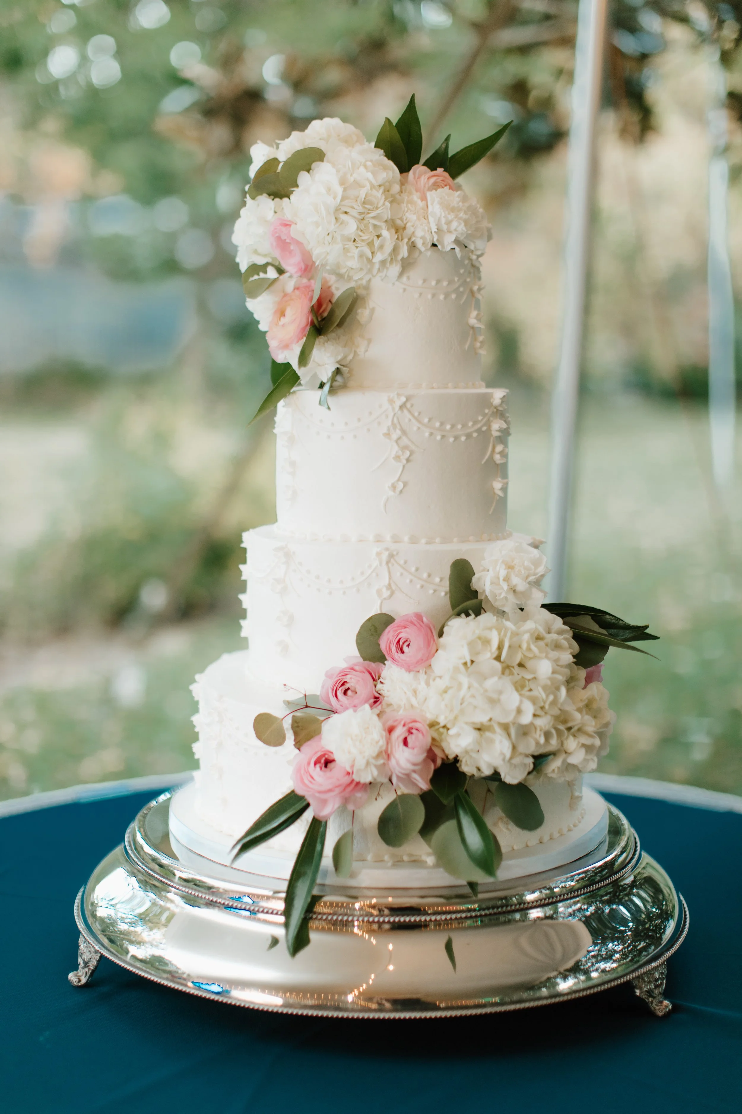 Three-tiered white wedding cake with floral decorations, including pink and white flowers and green leaves, on a silver stand.