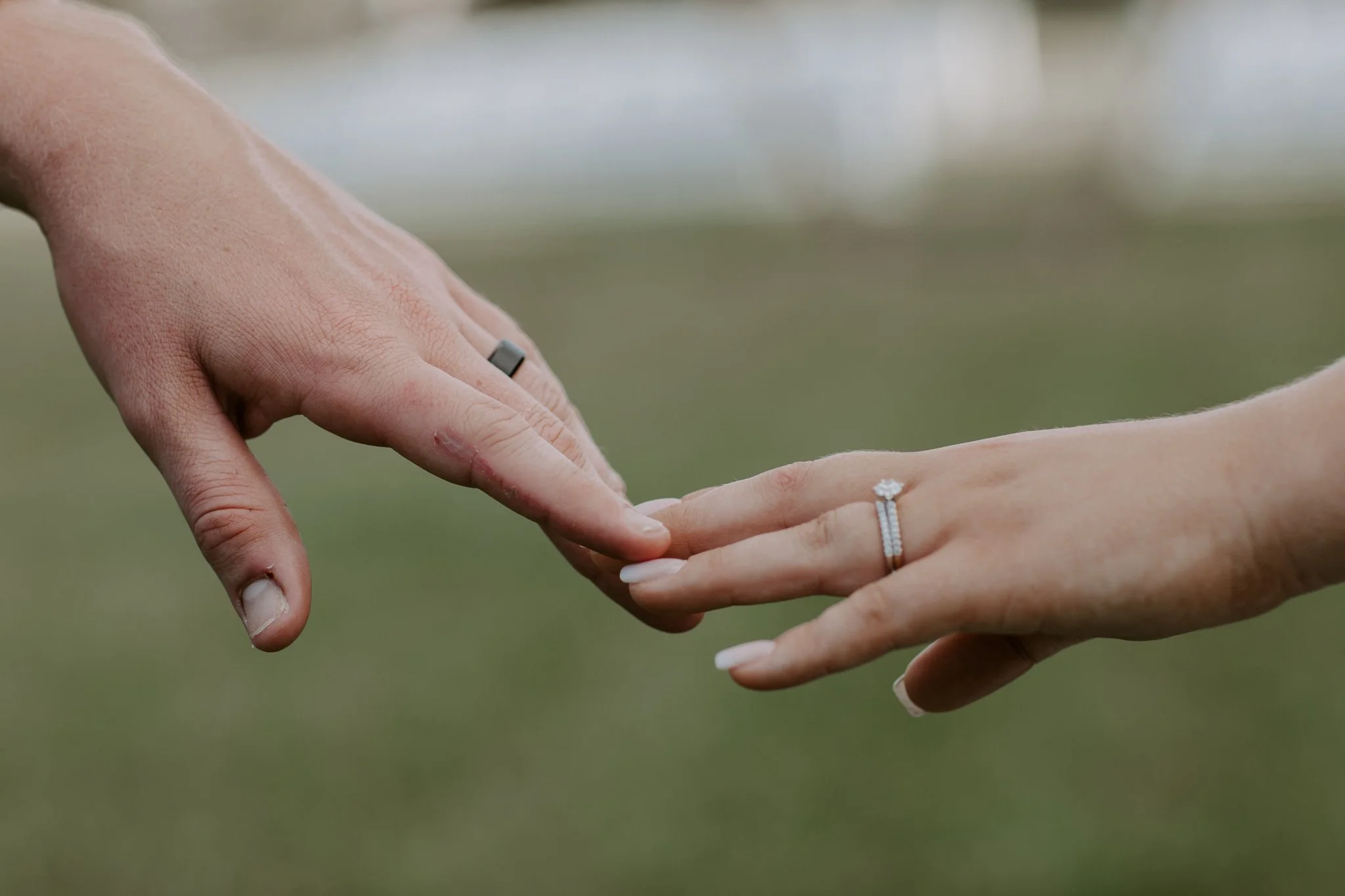 Close-up of two hands reaching towards each other, with one wearing a wedding ring and the other an engagement ring, against a blurred green background.