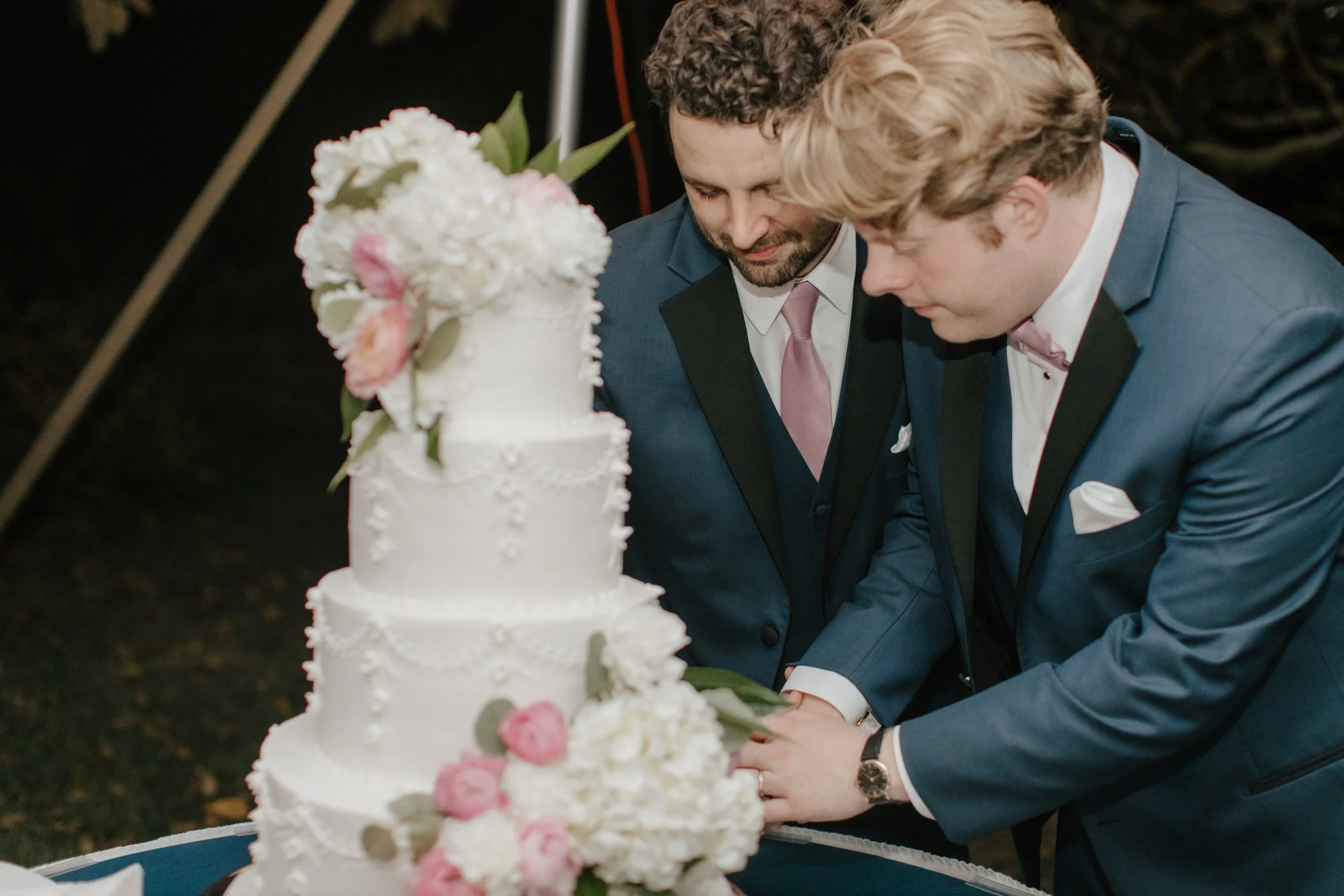 Two men in suits cutting a white tiered wedding cake decorated with flowers.
