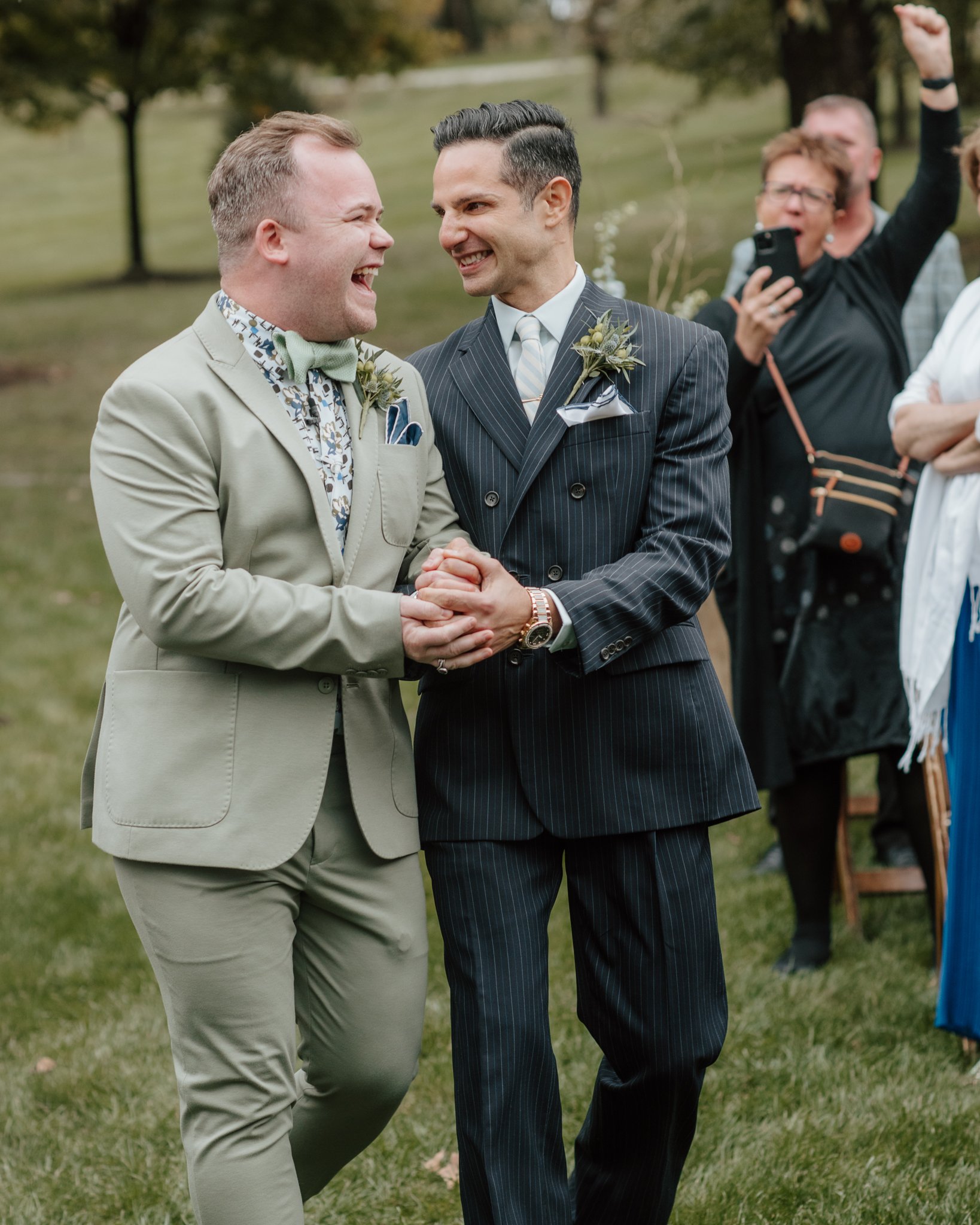 Two grooms celebrating and holding hands at an outdoor wedding ceremony with cheering guests in the background.