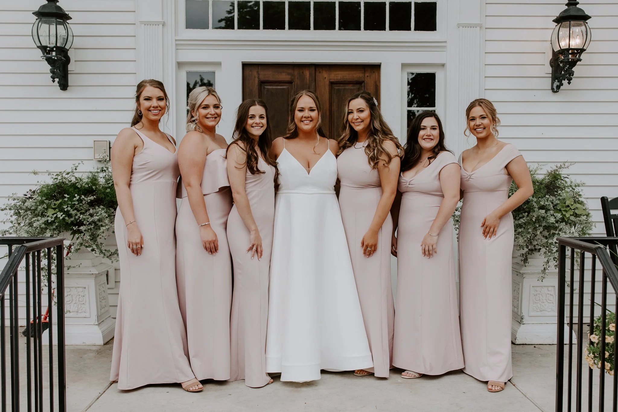 A bride standing with six bridesmaids in pale pink dresses, posing outside a white building with a wooden door and black lanterns.