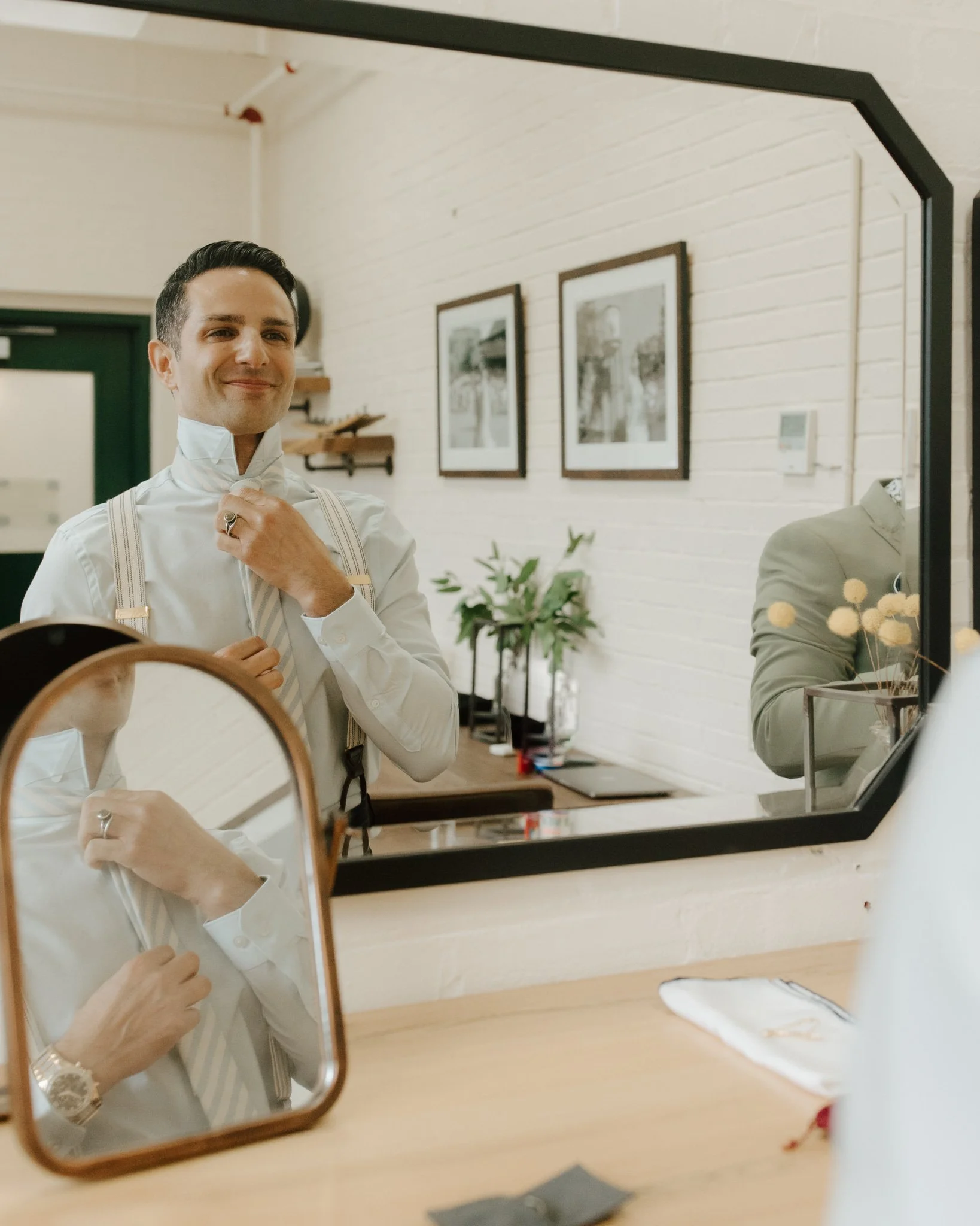 Man adjusting tie in front of mirror indoors with wall art and plants