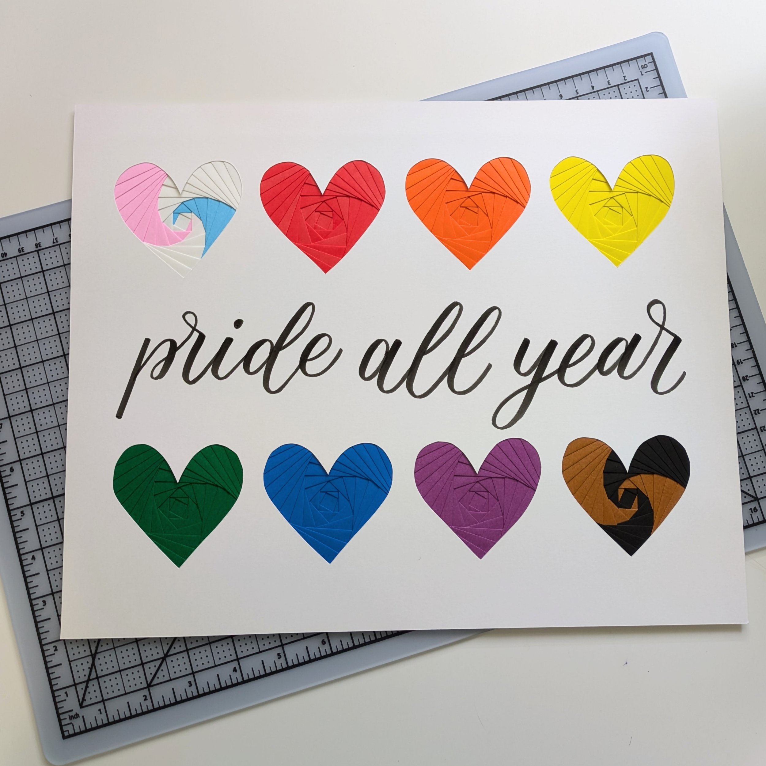 8 small iris folded hearts in progress pride flag colors with text "pride all year" in brush lettering