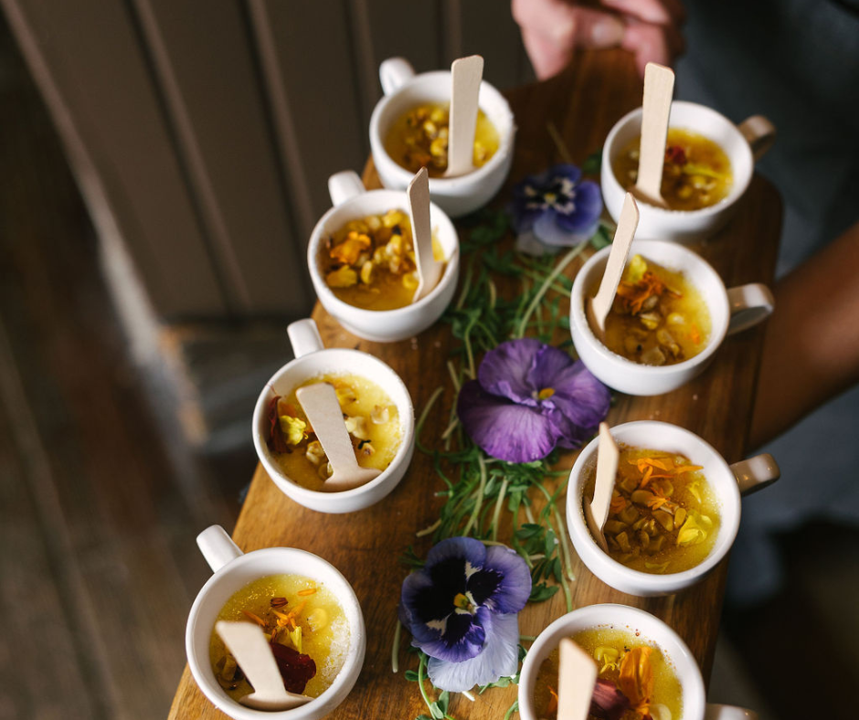 Nine small white cups filled with herbal tea, each with a wooden spoon inside, arranged on a wooden board decorated with purple and blue flowers and green leaves.