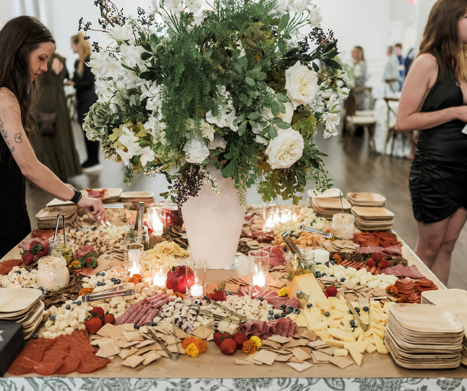 A large cheese and charcuterie platter with assorted cheeses, meats, berries, and crackers, topped with a tall floral arrangement in a white vase, on a decorated table at a social event.