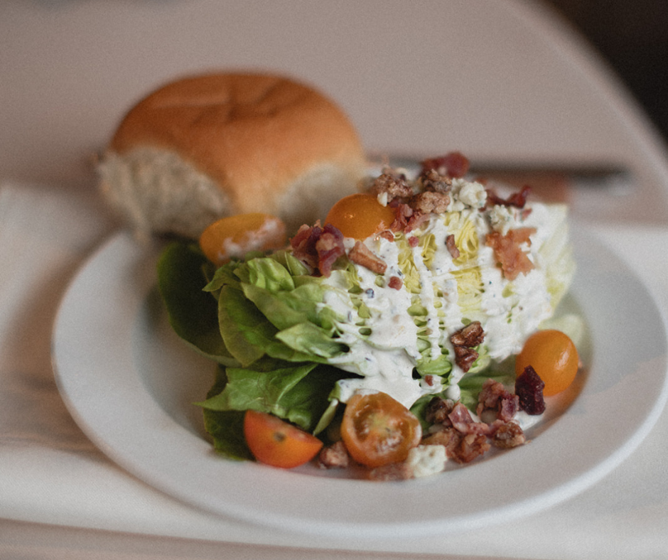 A plate of potato salad topped with bacon bits and cherry tomatoes, served with a bread roll.