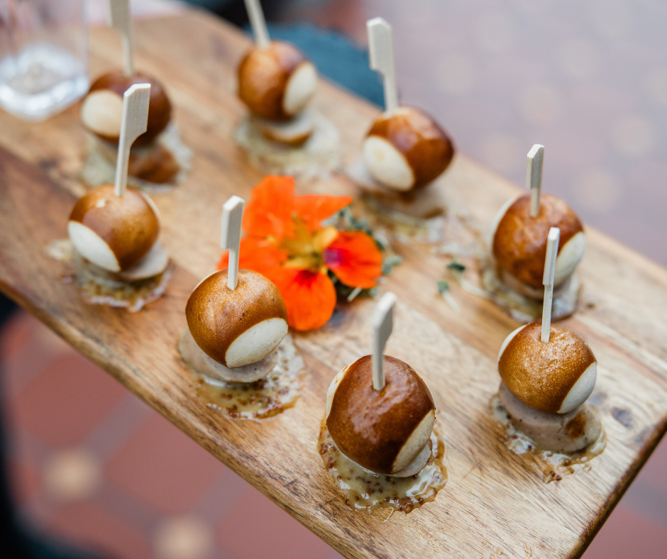 A wooden serving board with eight appetizers featuring small round pastries each topped with a white stick, garnished with orange edible flowers and green herbs.
