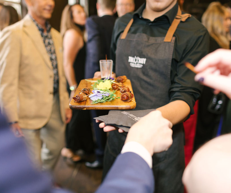 A waiter serving food on a wooden tray at a social event, with people in formal attire in the background.