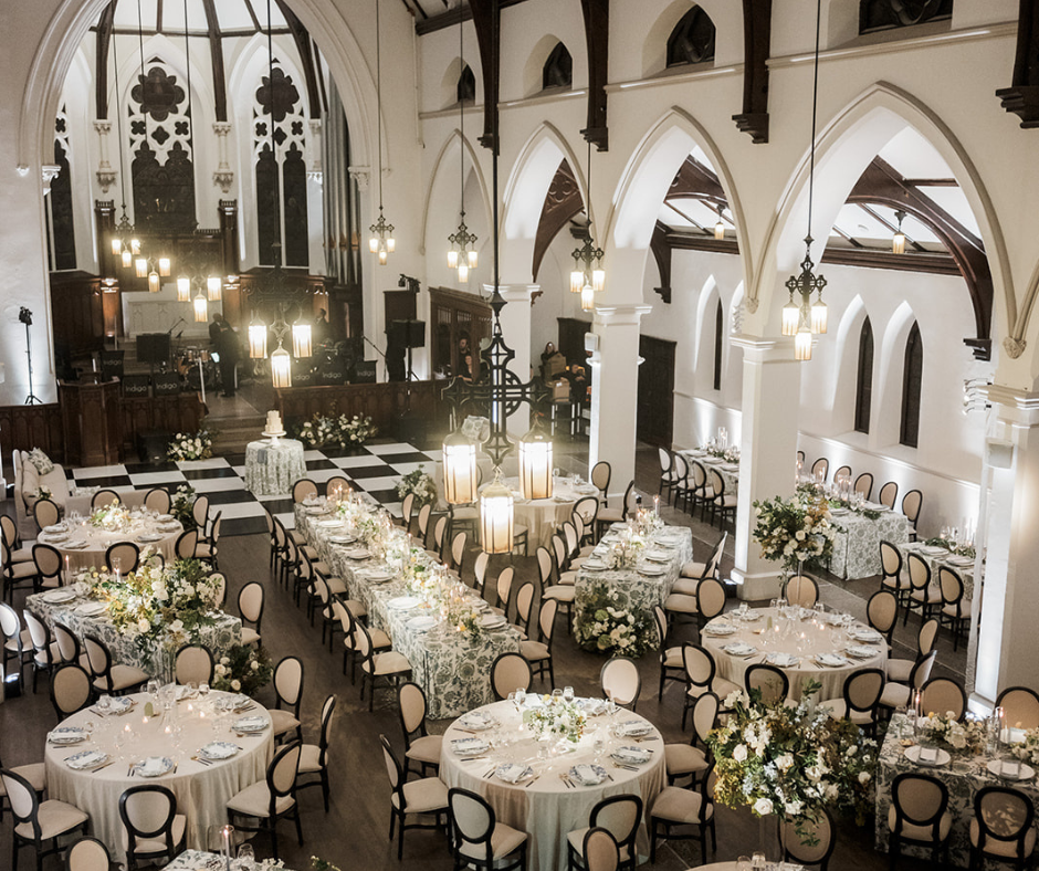 Elegant banquet hall decorated with floral centerpieces, round and rectangular tables with place settings, chandeliers, and Gothic-style arches and windows.