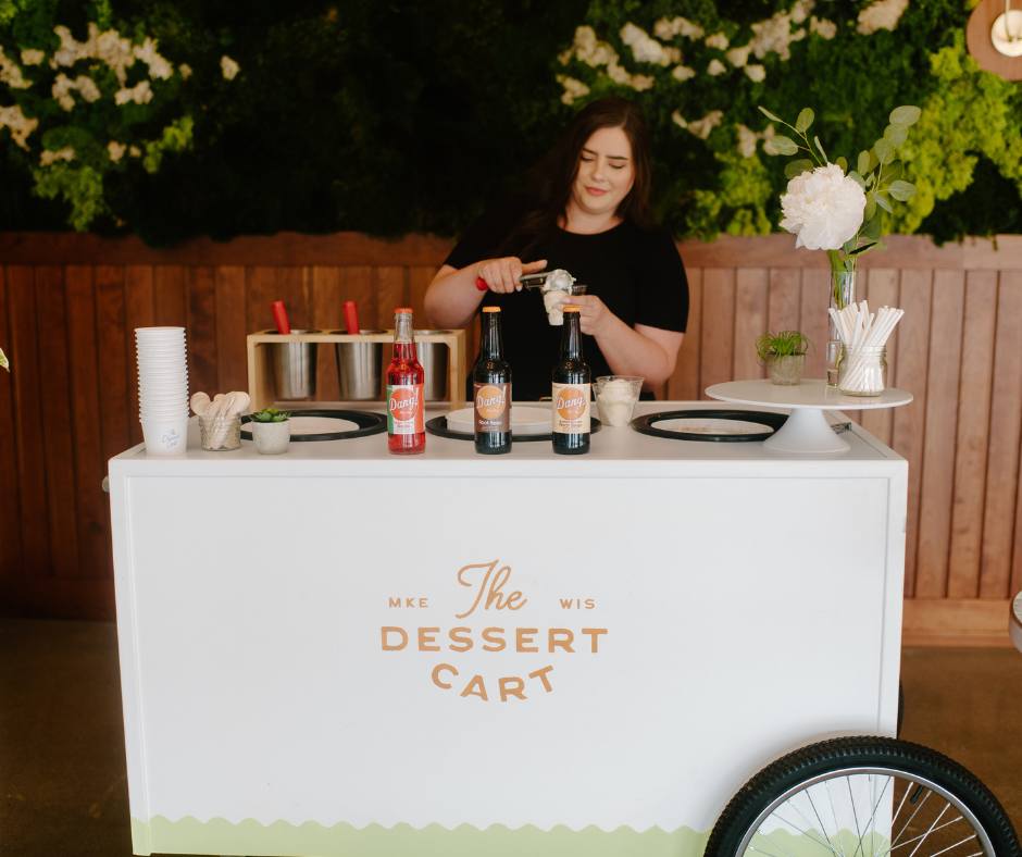 A woman making ice cream sundaes at a mobile dessert cart with flowers and beverages on display.