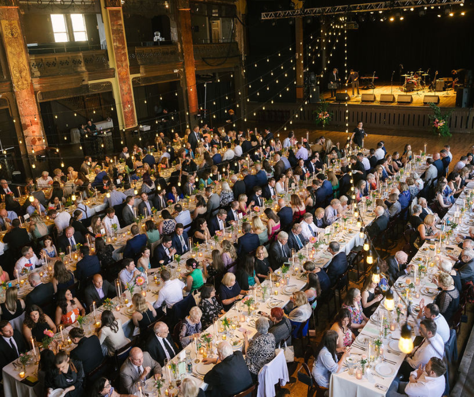 A large formal dinner event with hundreds of people seated at long decorated tables, a stage with musicians, and string lights hanging above in a historic venue.