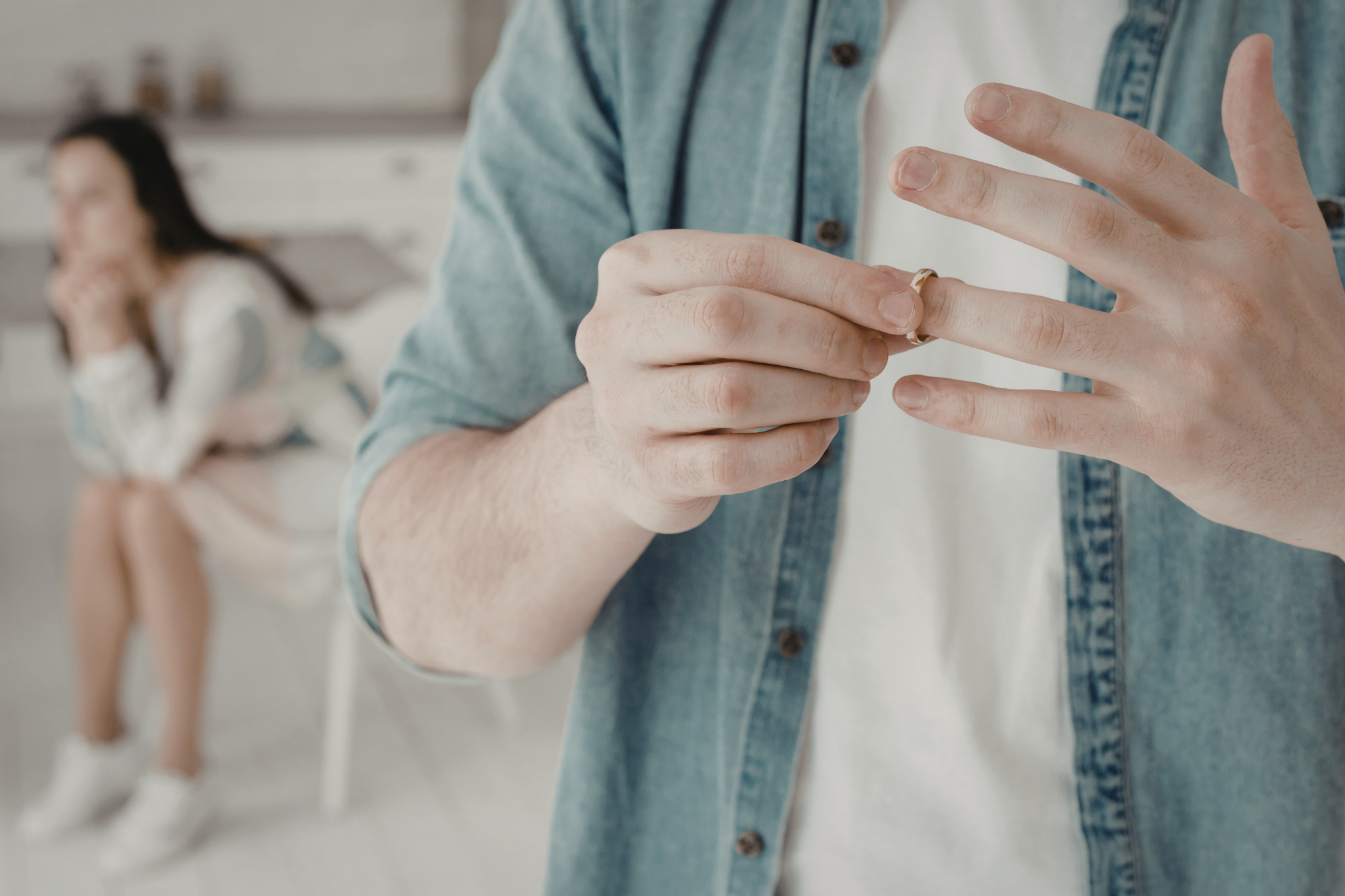 A man removes his wedding ring while his partner sits blurred in the background. Healing after betrayal is a process. Find compassionate affair recovery counseling to help you navigate this pain in Portland, Oregon, zip code 97210.