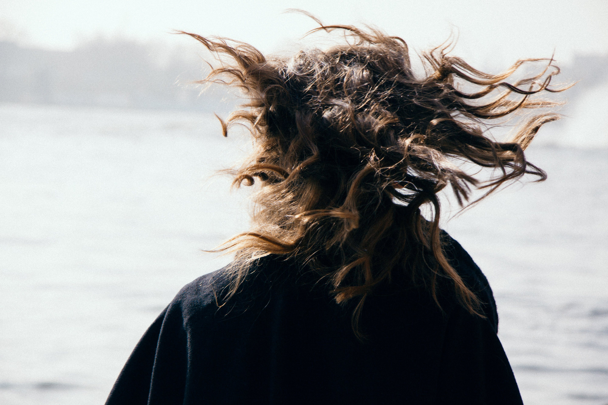 A person with long, curly hair being swept by the wind while standing near a body of water in Portland, Oregon, or Seattle, Washington, symbolizing the emotional turbulence and longing for connection often experienced in anxious attachment