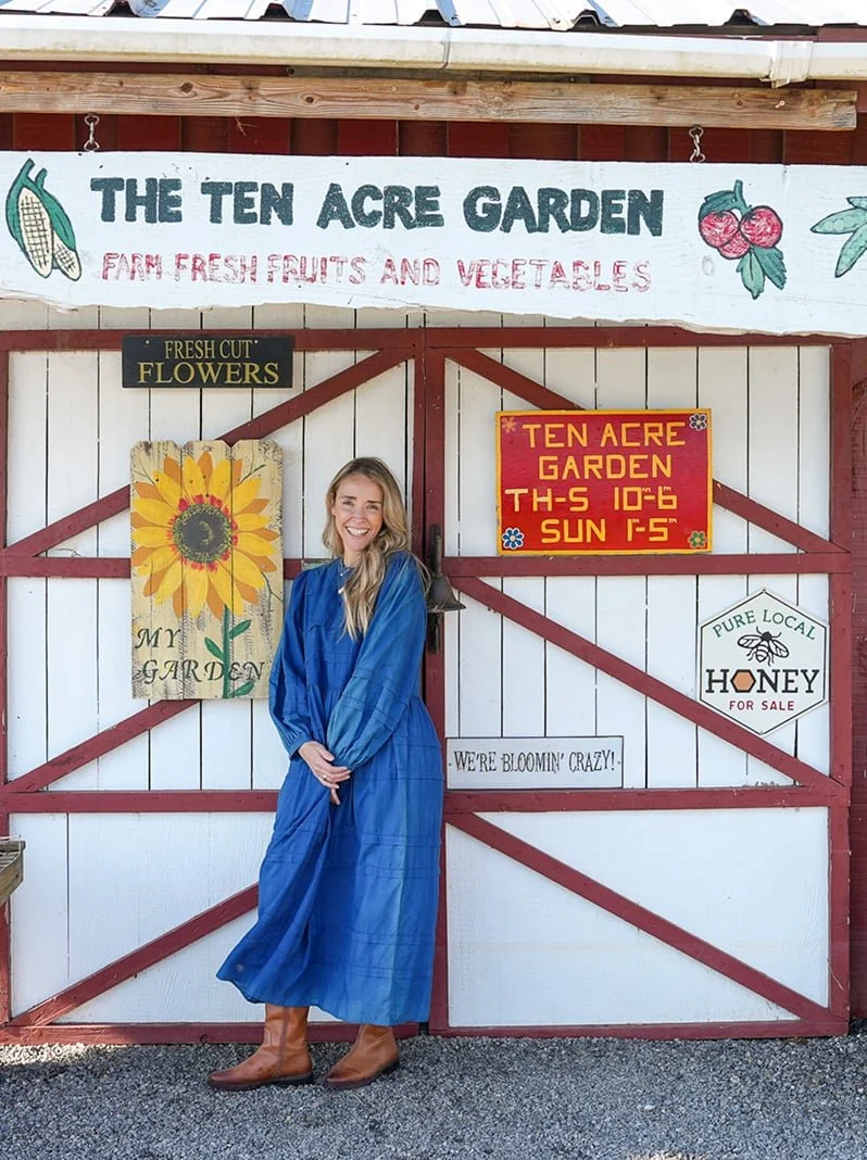 A spiritual mentor in a blue dress stands smiling at The Ten Acre Garden produce stand with colorful signs and a sunflower mural.