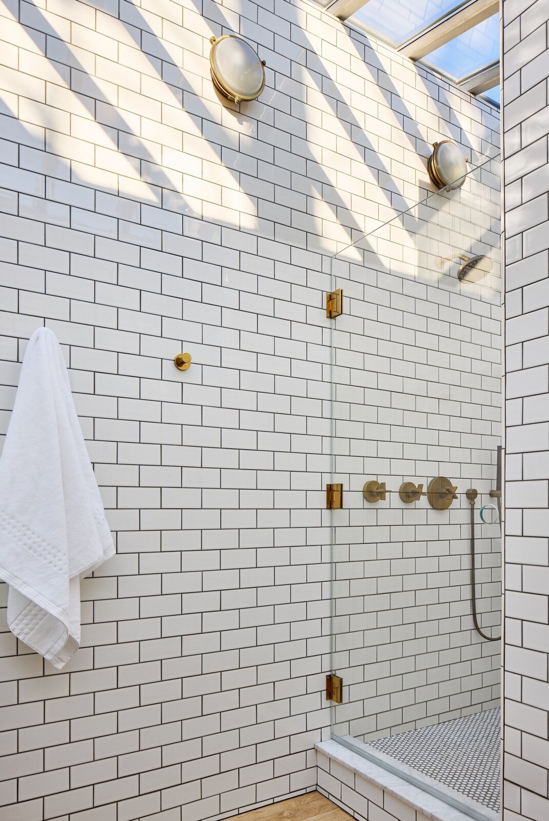  White subway tile with charcoal-colored grout covers the primary bath where sunlight streams in from overhead thanks to a huge, new skylight that is 10 feet at its highest point. Wall sconces from The Urban Electric Co. lend a nautical feel. 