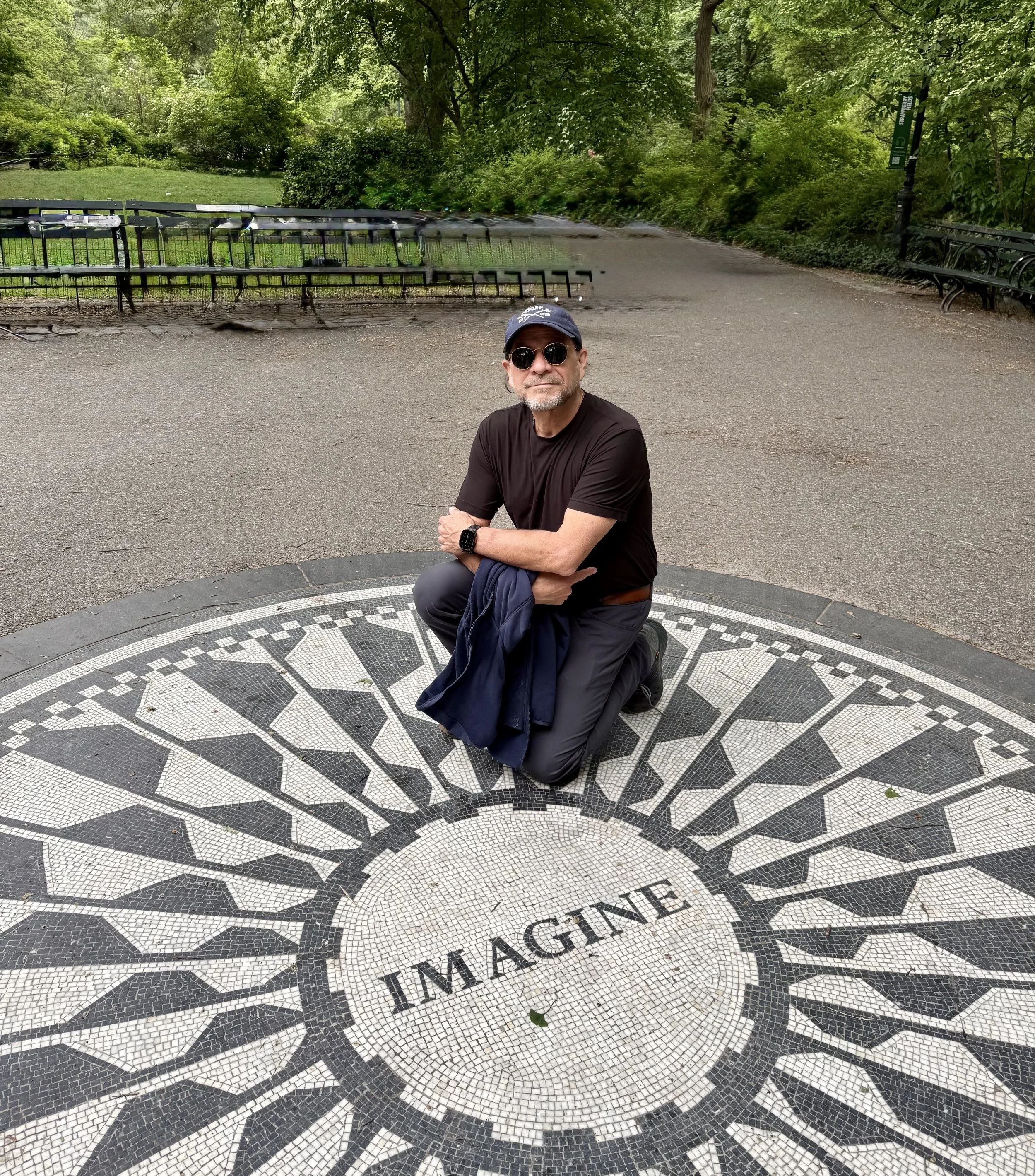 A man with a beard, wearing sunglasses, a black cap, and black clothing, kneels on a black and white mosaic circle in a park surrounded by greenery and benches, with a distorted bench and a lamppost in the background.