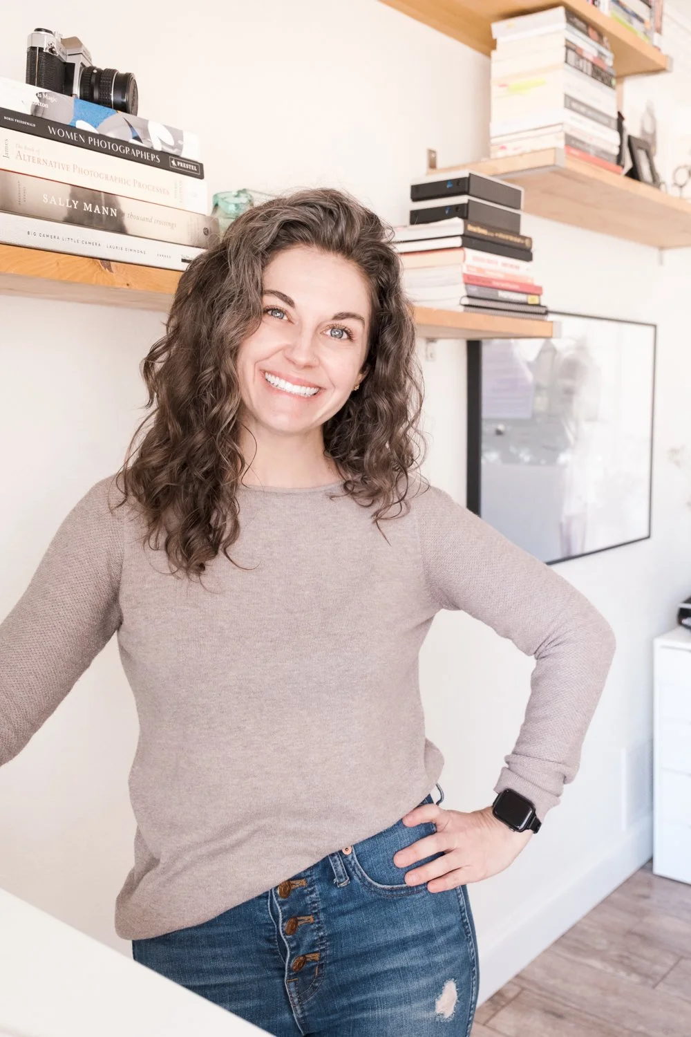 Founder of Made of Gray Fort Worth Web Design. White women with curly hair smiling at the camera in her studio with shelves and books in the background.
