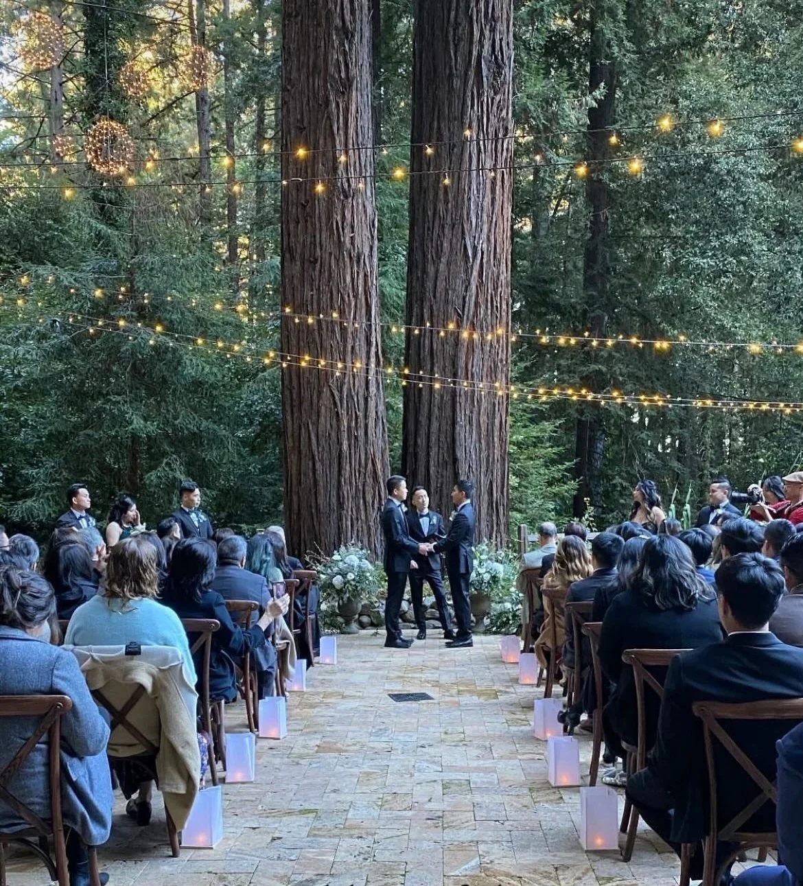 Grooms saying their vows under a pair of redwood trees, guests seated, on a travertine patio