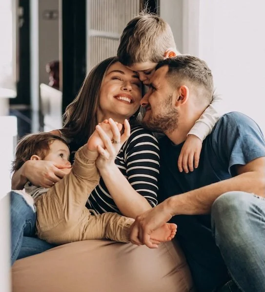 Family of four happily hugging on a couch, with a woman and man smiling, and two children, one hugging the woman and the other leaning over the man.