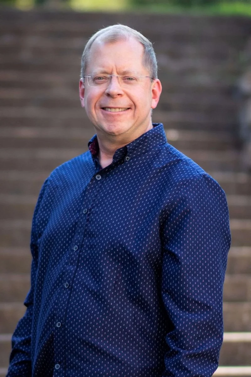 A man with glasses wearing a navy blue shirt with small white polka dots, standing outdoors with a blurred wooden staircase in the background.