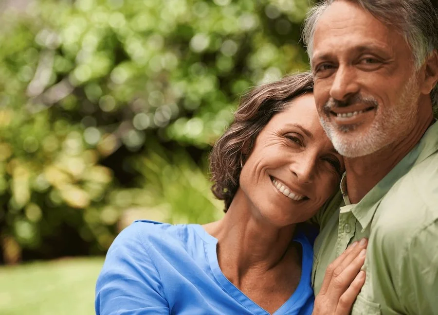 A smiling middle-aged couple hugging outdoors in a green, leafy setting.