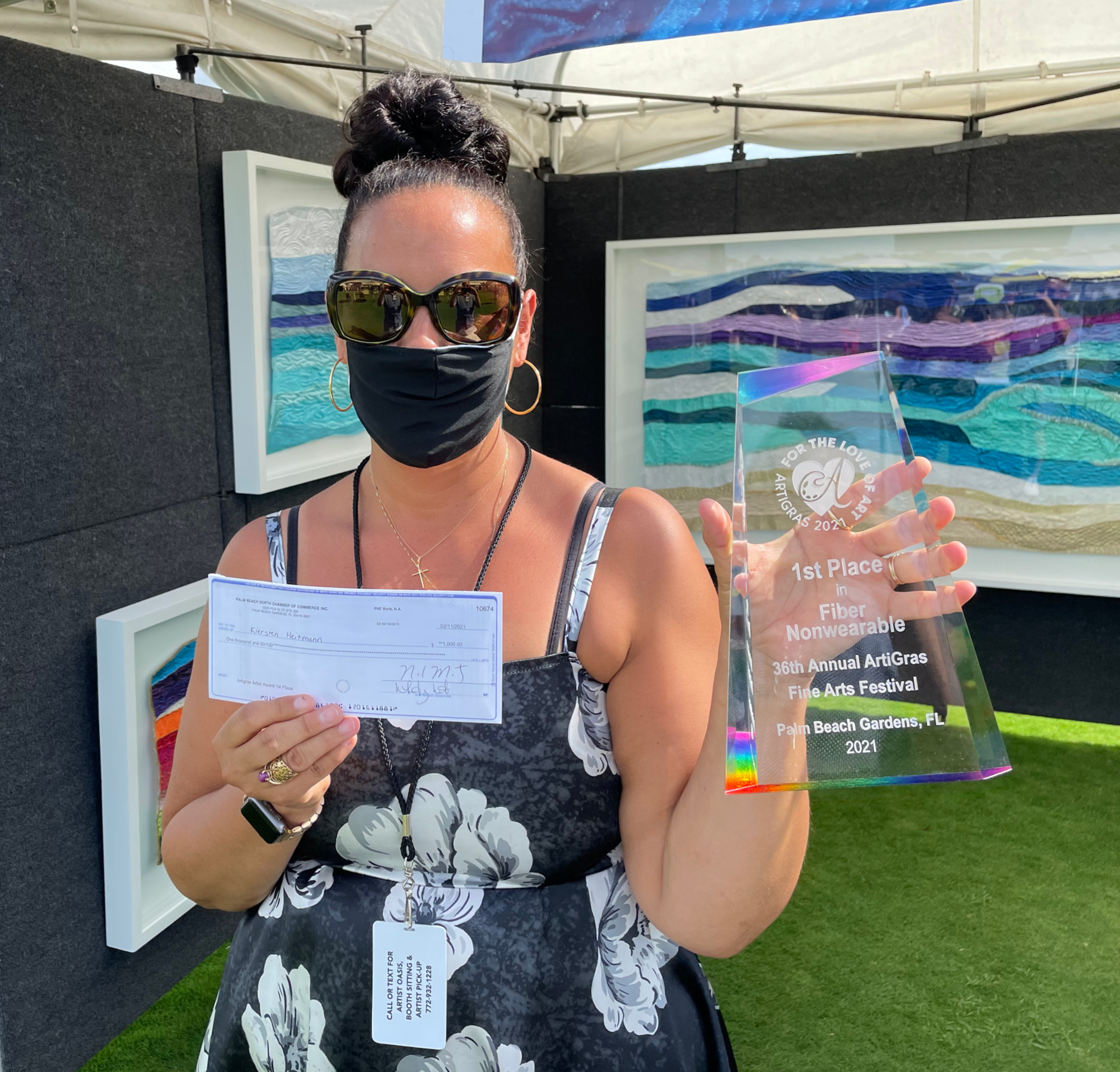 A woman in a floral dress with sunglasses and gold hoop earrings, wearing a black face mask, is holding a check in one hand and a crystal award plaque in the other. The award reads '1st Place in Fiber , 36th Annual ArtiGras Fine Arts Festival, Palm Beach Gardens, FL, 2021'.