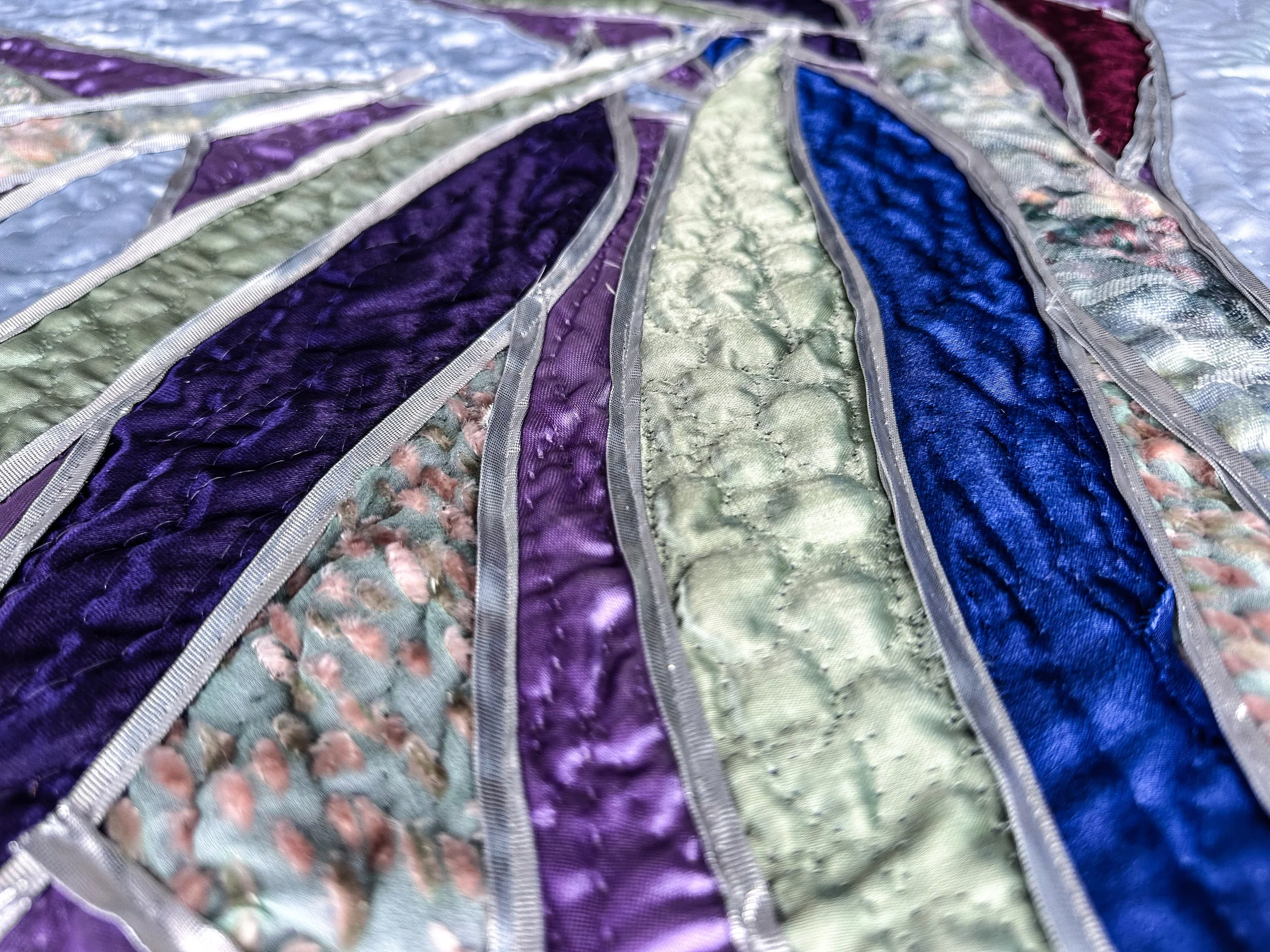 Close-up of a colorful fiber art piece with vertical strips of purple, blue, green, and patterned designs, edged with silver trim.