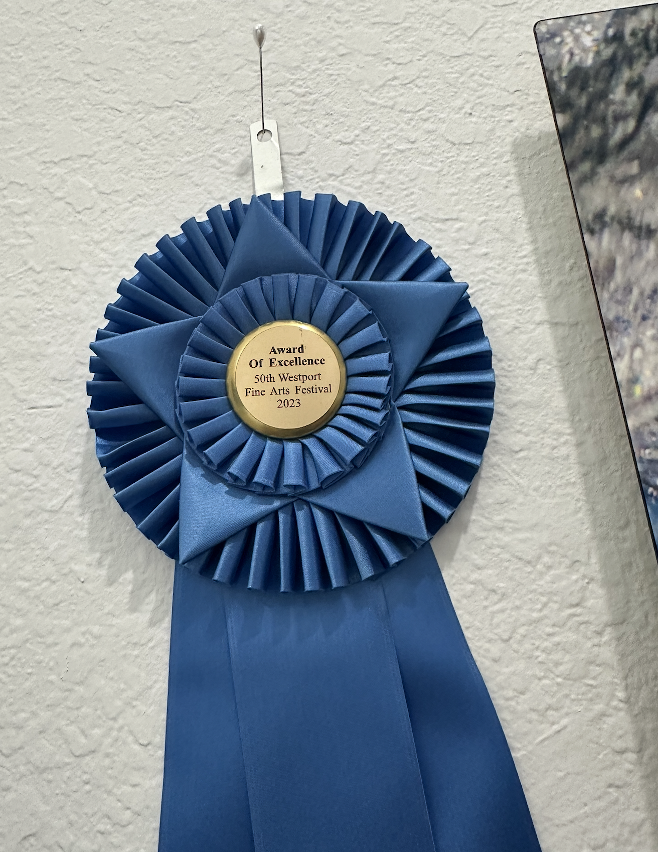 Blue award rosette ribbon with a central gold medallion reading "Award of Excellence, 50th Westport Fine Arts Festival 2023," hanging on a light-colored wall.