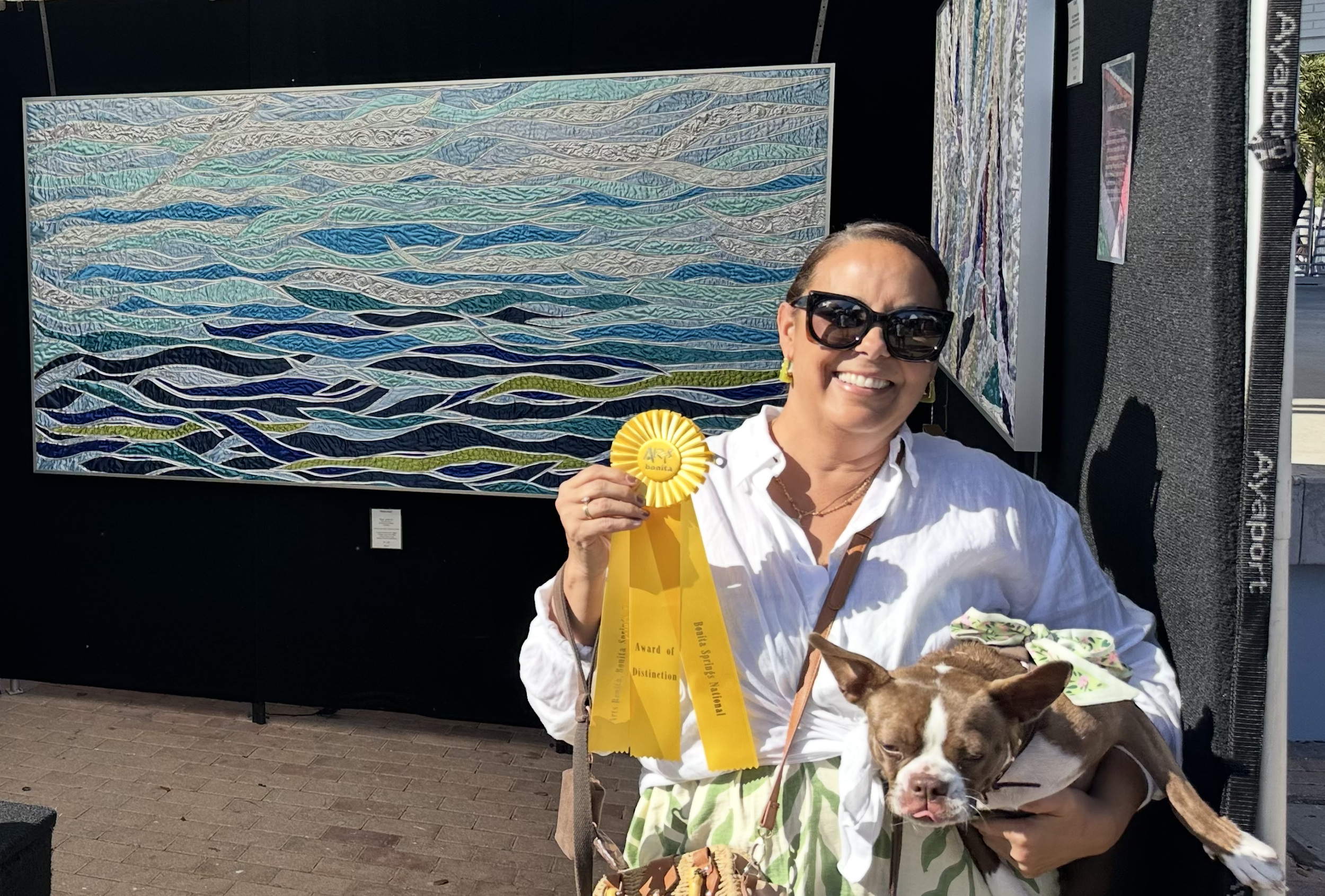 Woman smiling and wearing sunglasses, holding a yellow award ribbon and a small dog, in front of abstract artwork at an outdoor art event.