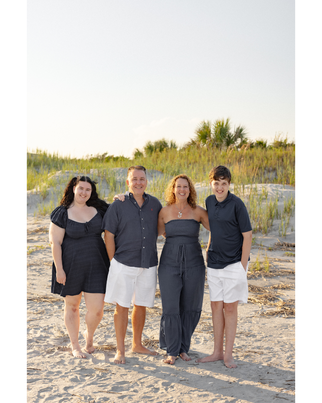 Four people standing on a sandy beach, smiling, with grassy dunes and a few trees in the background, during daytime.
