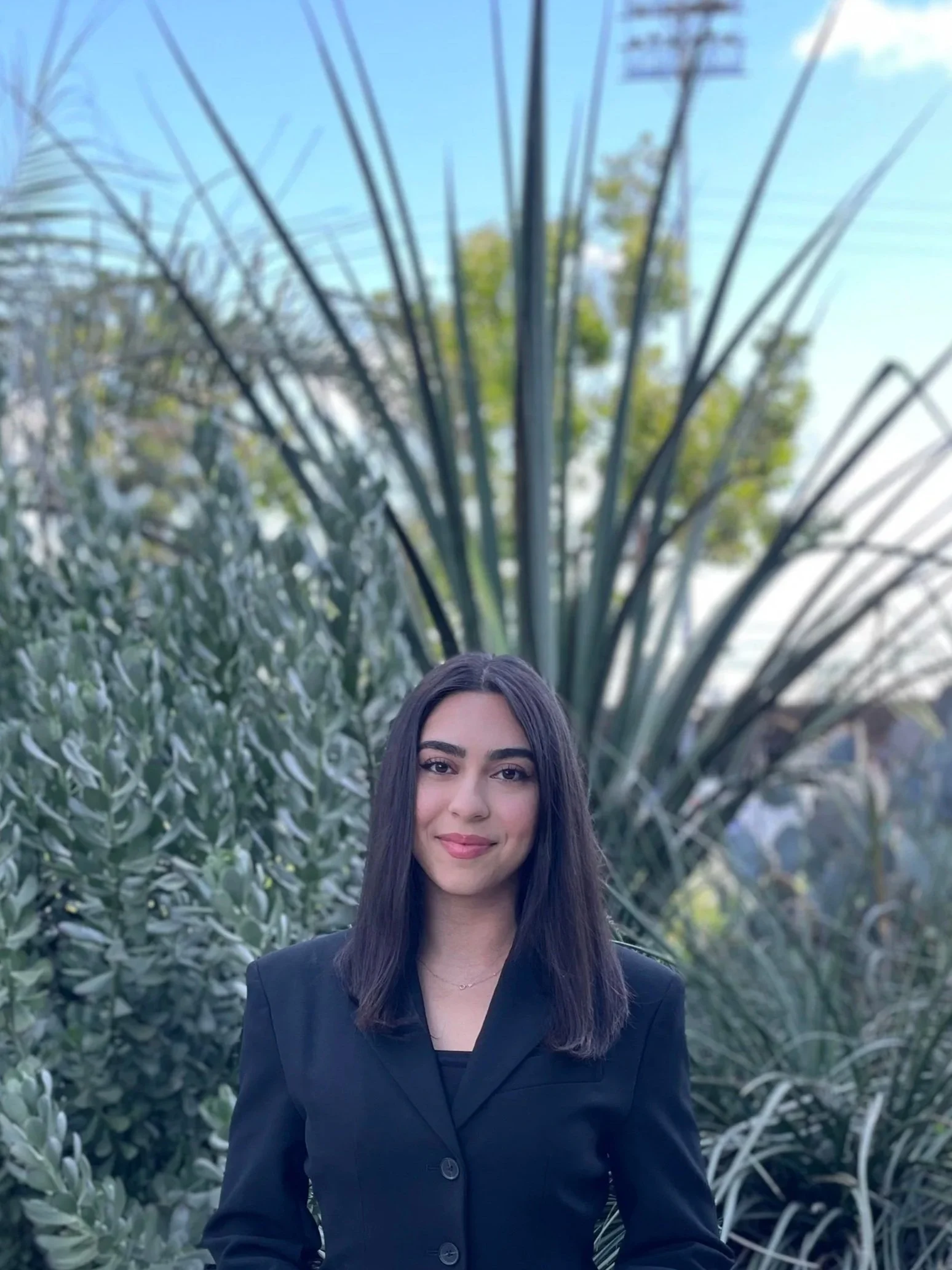 A woman with long dark hair and a friendly expression, wearing a black blazer, standing outdoors in front of large, spiky green plants against a blue sky.