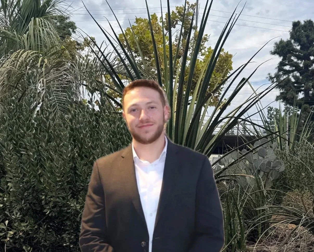 A young man in a black suit and white shirt standing outdoors in front of lush green tropical plants.