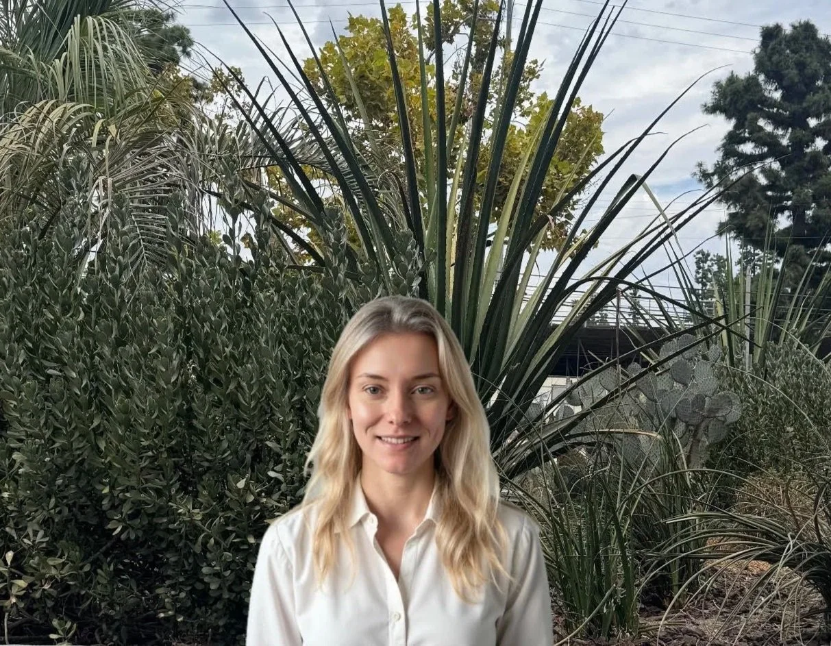 A woman with blonde wavy hair wearing a light-colored blouse standing outdoors in front of various large green plants and trees.