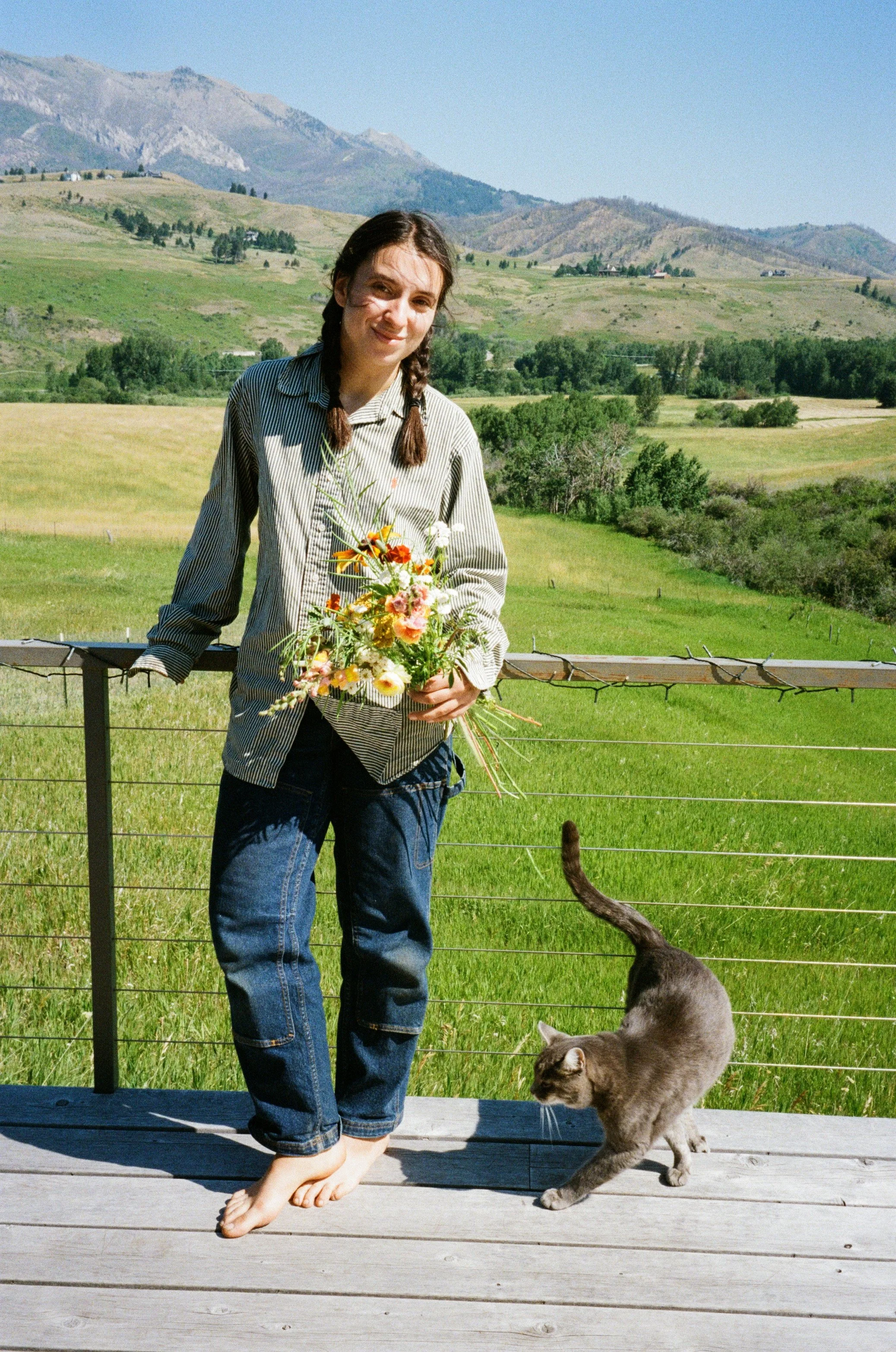A woman holding a bouquet of flowers standing on a wooden deck with a gray cat nearby, overlooking green fields and mountains under a clear sky.
