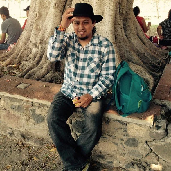 Joven con sombrero negro, camiseta a cuadros azul y blanca, sentado en un muro de piedra, junto a un árbol grande, mochila junto a él, en un parque.