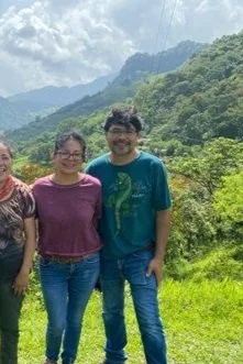 Tres personas de pie en un campo verde con montañas y árboles en el fondo, cielo parcialmente nublado.