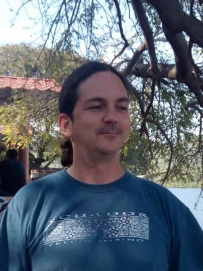 Hombre de cabello largo y camisa azul oscuro, de fondo árboles y un lago o río, en un entorno natural.