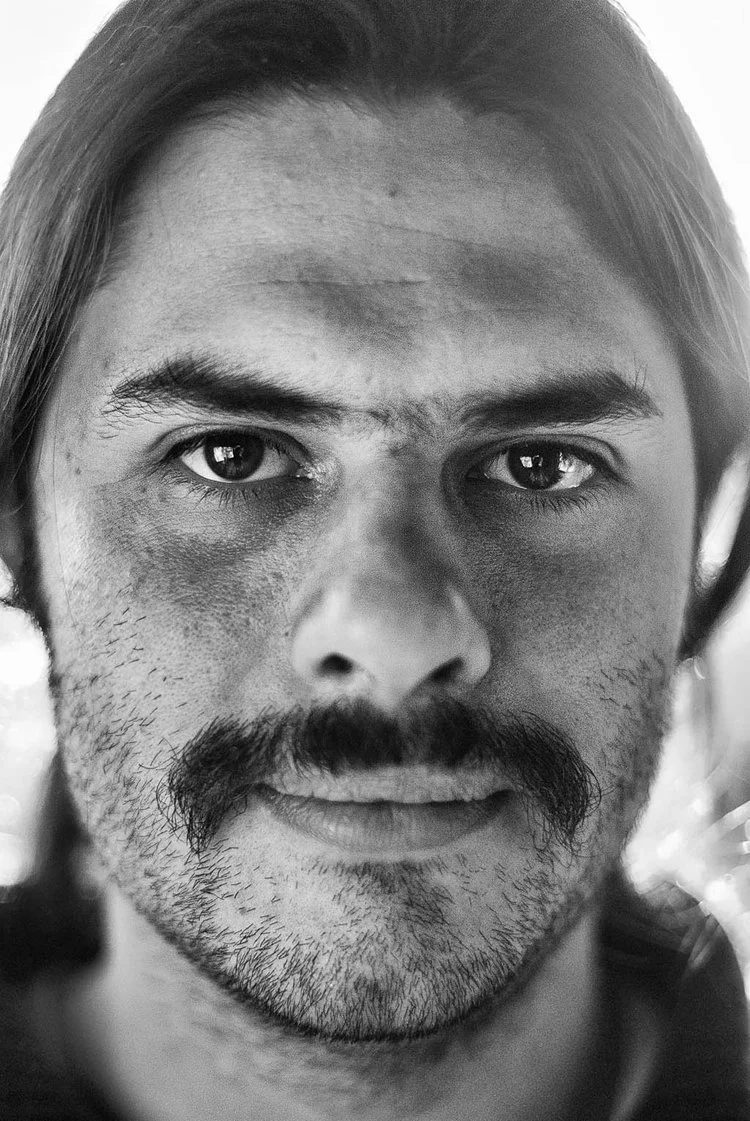 Retrato en blanco y negro de un hombre con barba y bigote, ojos claros y cabello largo, mirando directamente a la cámara.