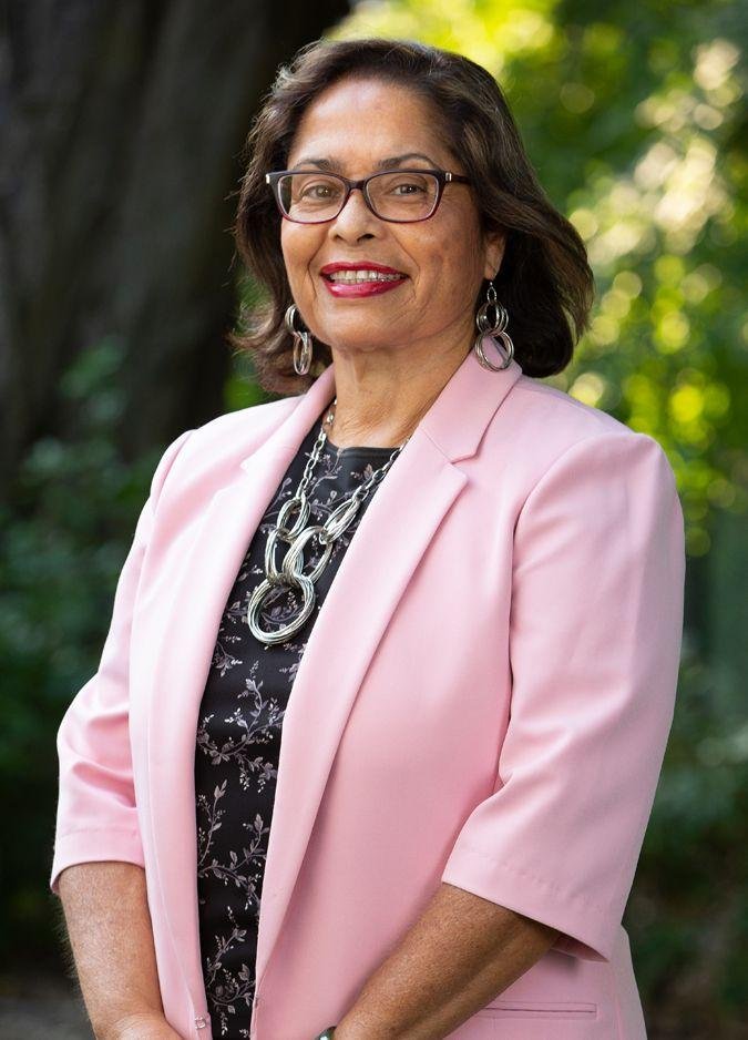 Mujer mayor sonriendo, lleva gafas, chaqueta rosa y accesorios plateados, en un entorno al aire libre con árboles y luz natural.