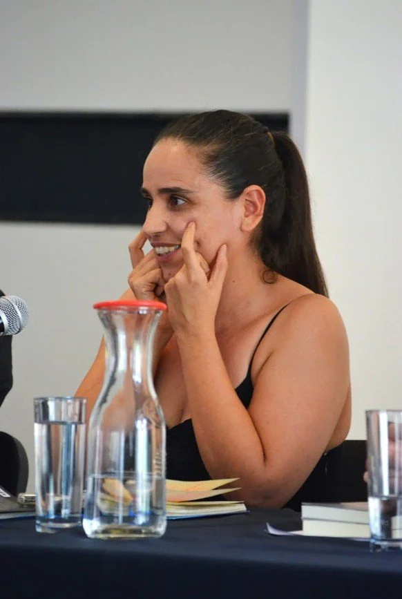 Una mujer con cabello recogido, sonriendo, con las manos en el rostro, en una mesa con libros, agua y un micrófono.
