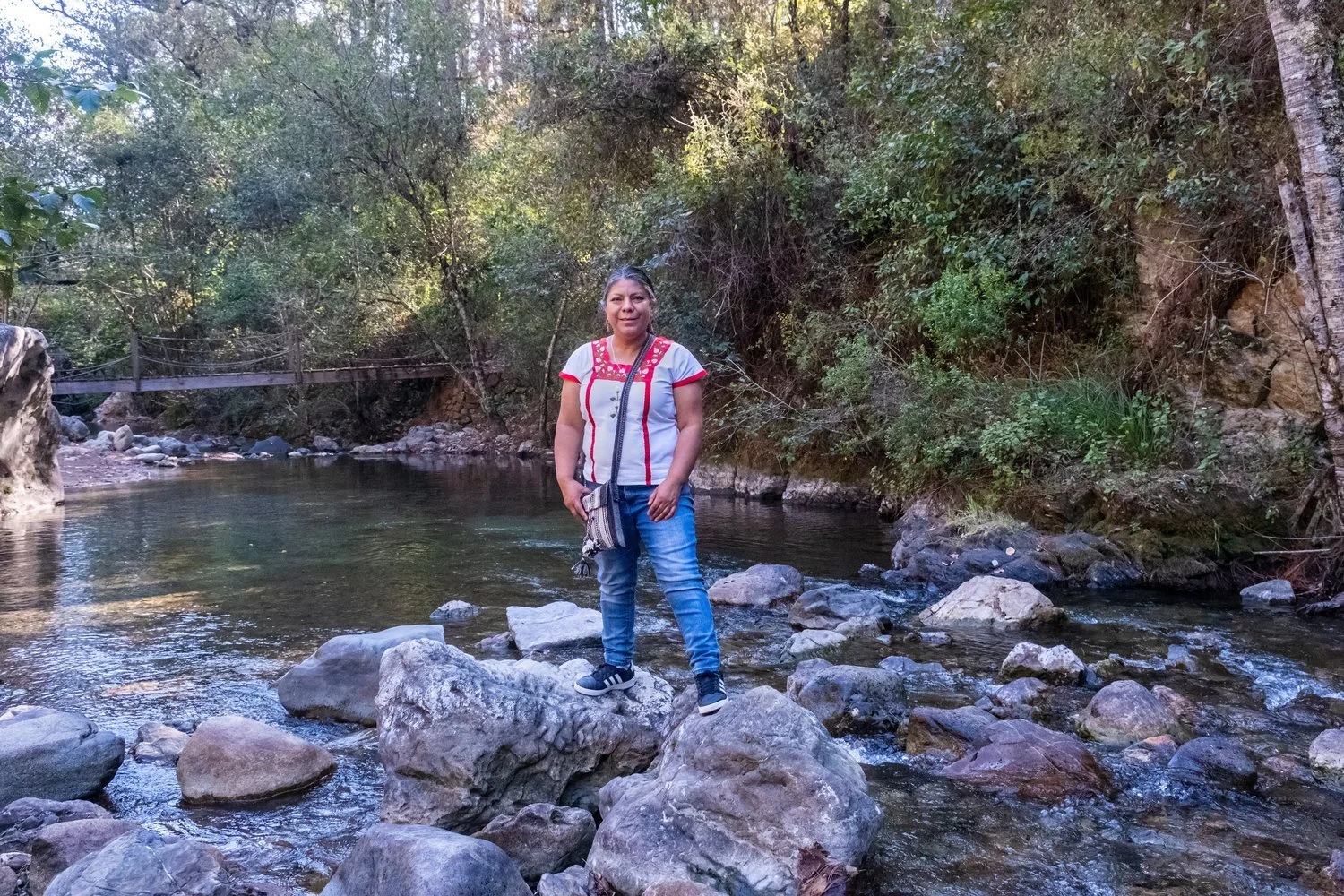 Mujer en pantalon y camiseta blanca con detalles rojos, de cabello oscuro, en un río con rocas y árboles verdes en un entorno natural.