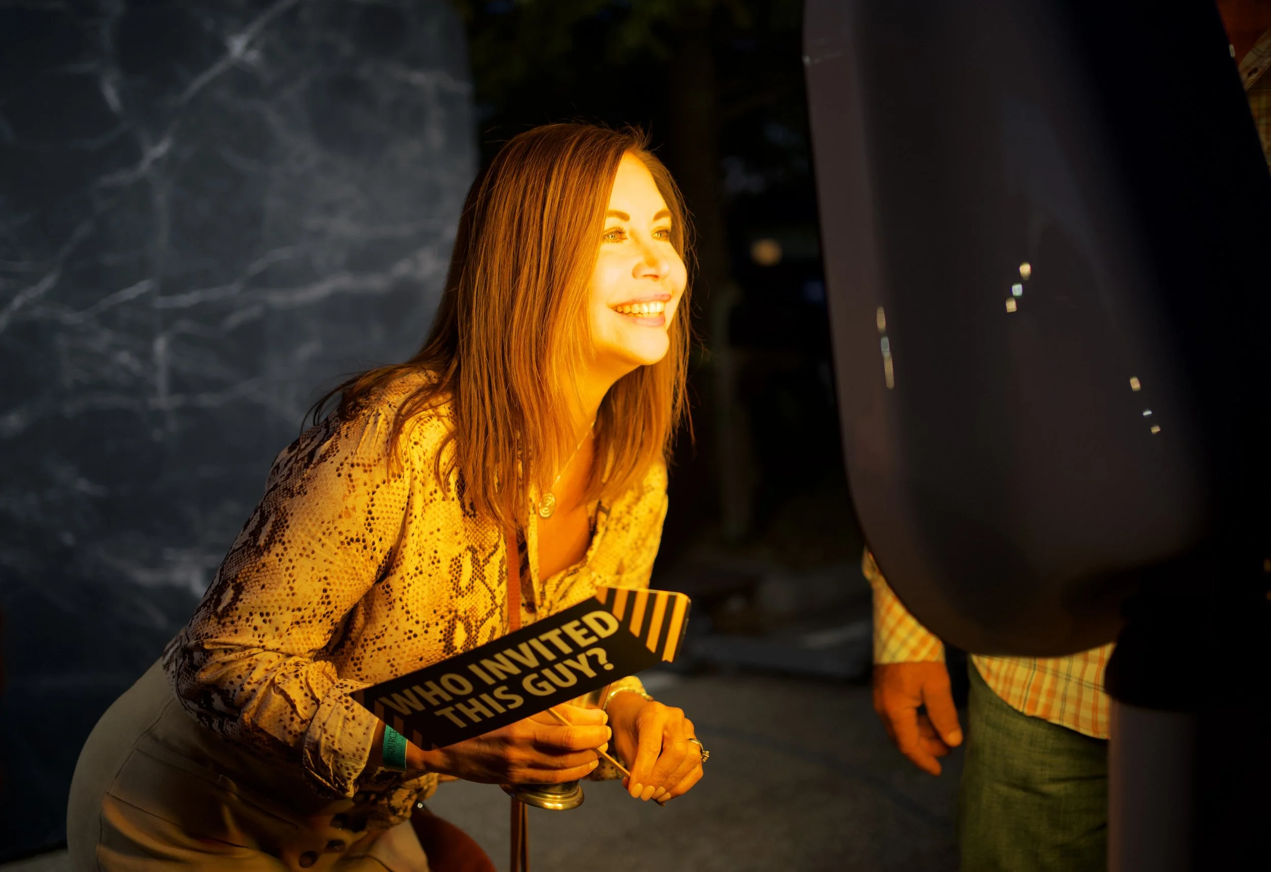 A woman with long brown hair smiling while holding a sign that says 'Who invited this guy?'. She is dressed in a patterned yellow top and tan pants, and is engaged in conversation or interview, with equipment or crew partially visible around her.