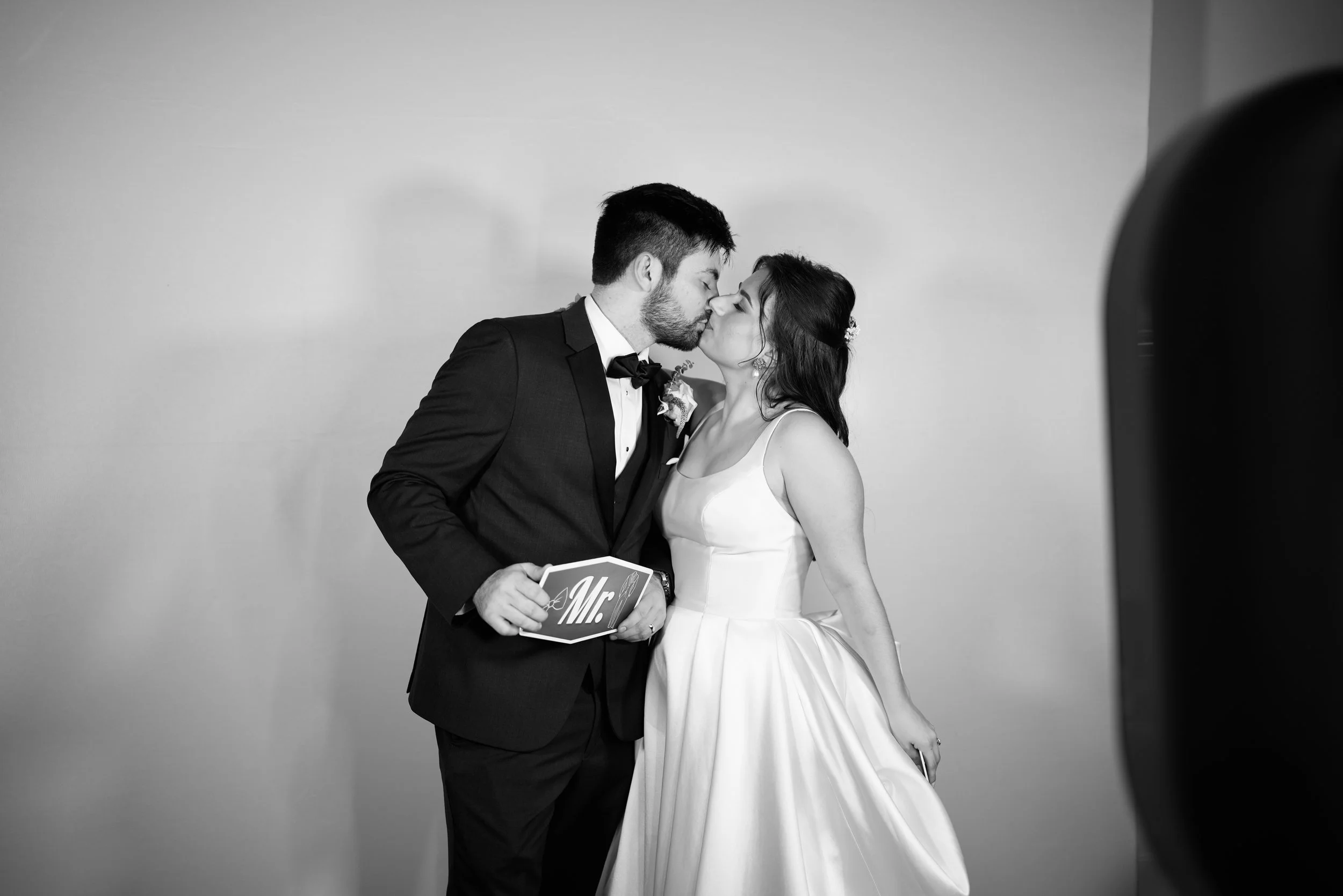 A black and white photo of a couple dressed in wedding attire sharing a kiss. The groom is in a tuxedo with a bow tie, holding a small book or card that says "Mr." The bride is in a white gown, slightly lifting the skirt.