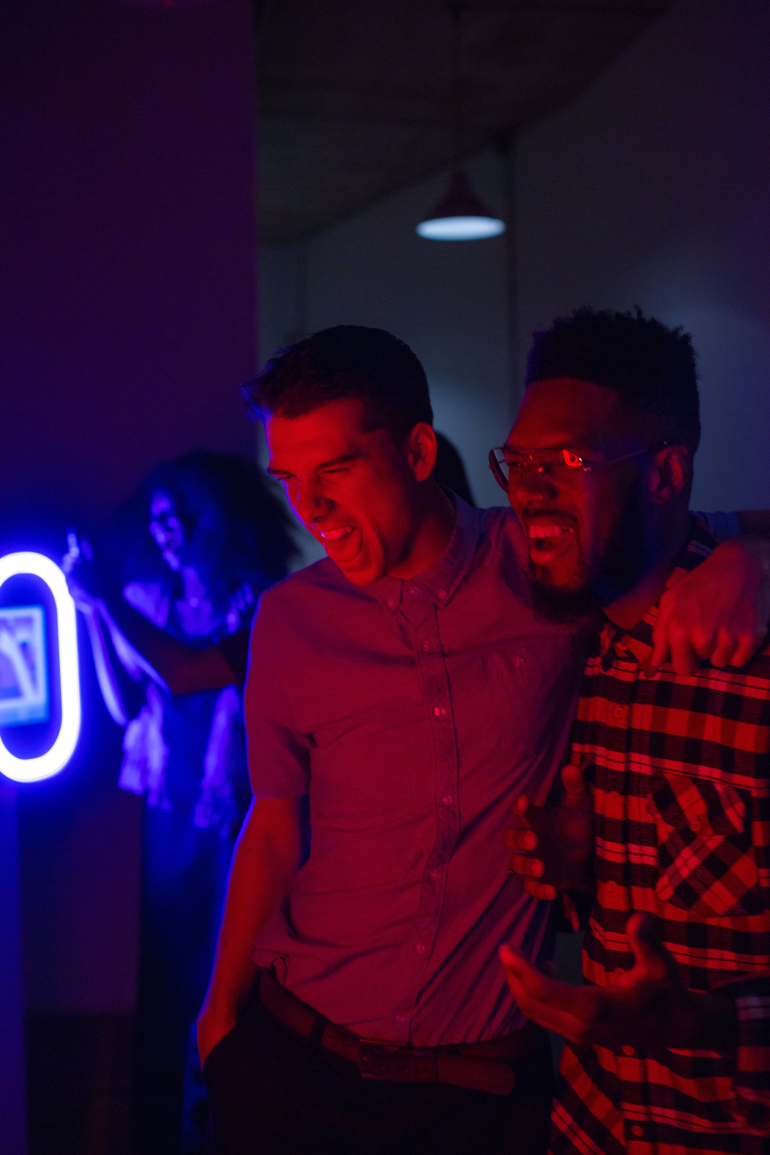 Two young men are enjoying and laughing together at a nightclub or bar, with colorful neon lighting illuminating the background.