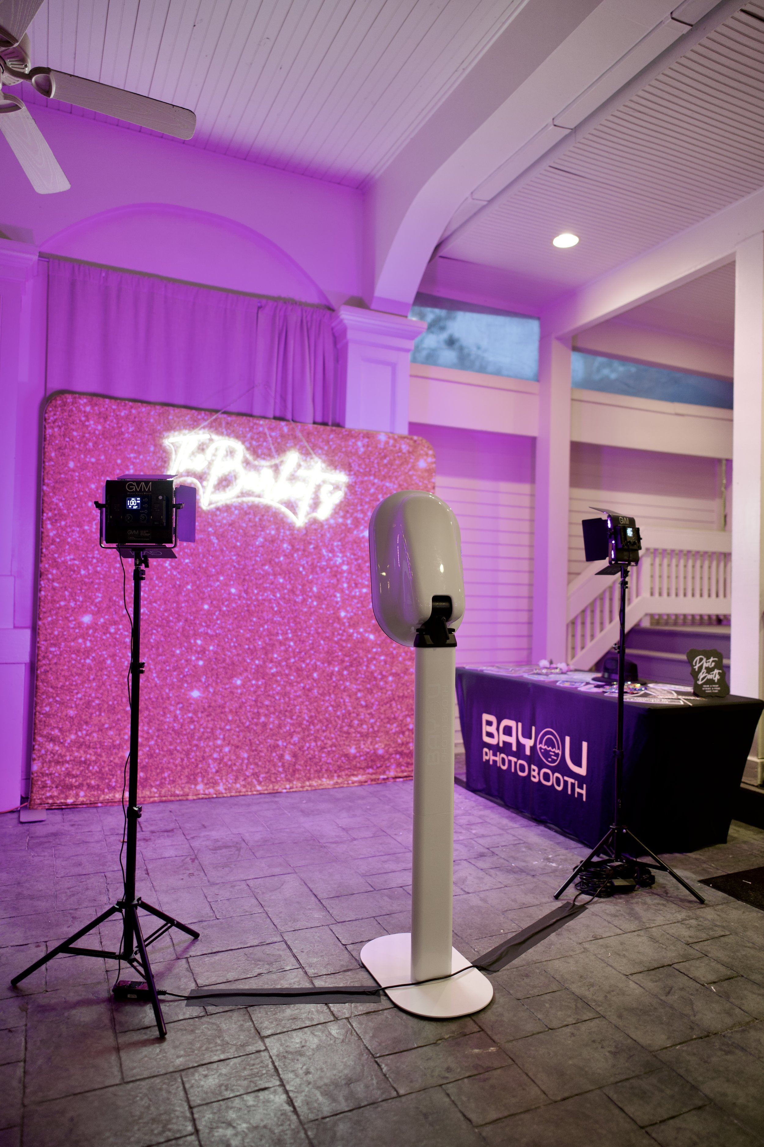 A photo booth setup with a pink glittery backdrop, two professional lights, and a white automatic sanitizer dispenser in front. A table labeled 'Bayou Photo Booth' is on the right side.