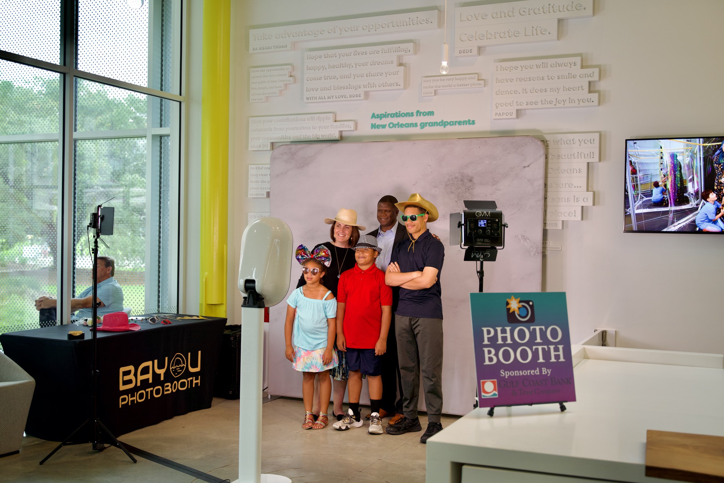 People posing for a photo at a photo booth in an indoor space with a large window showing greenery outside. There are four individuals, two children and two adults, wearing hats and sunglasses. The photo booth is marked by a sign on a table that read