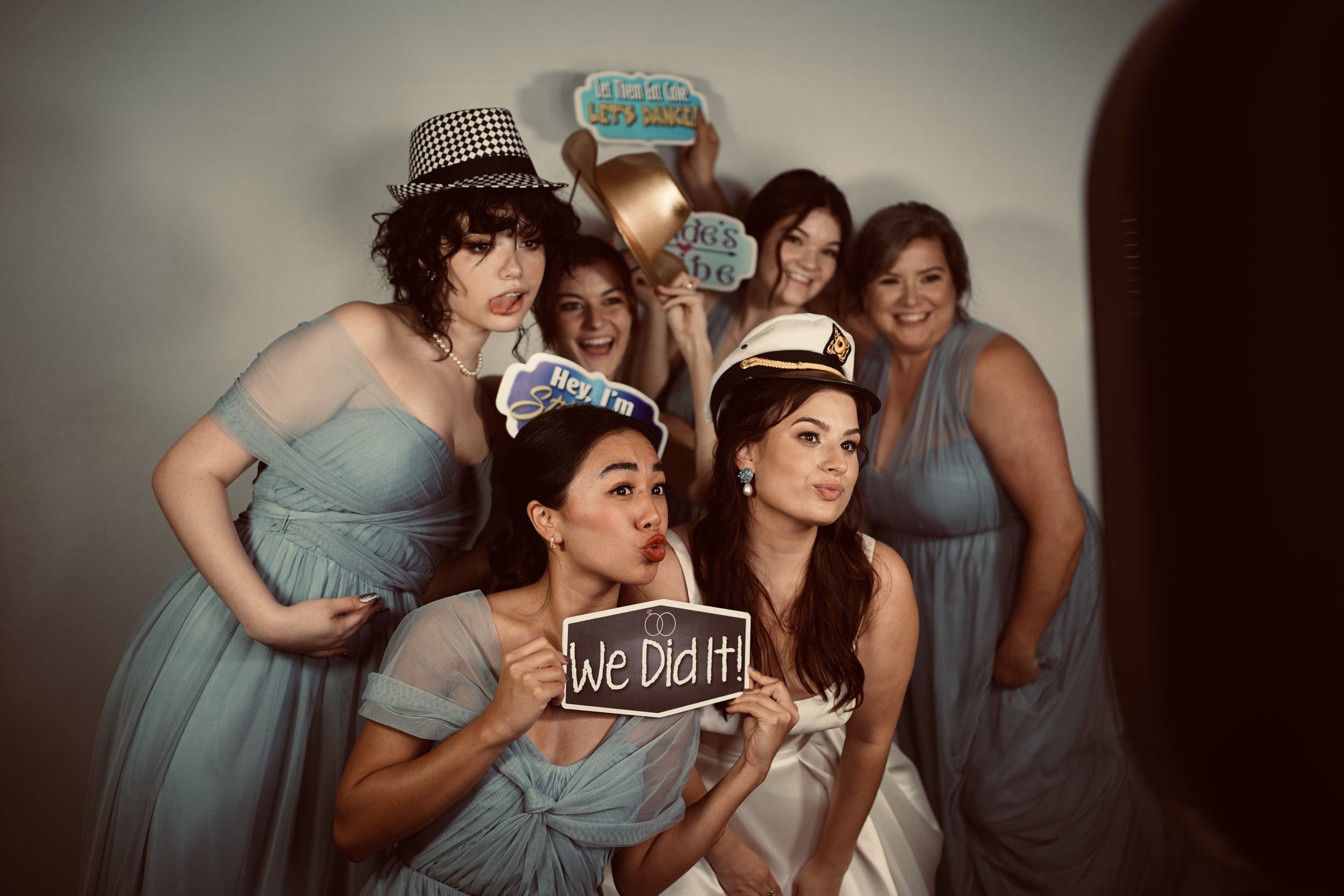 Group of women dressed in blue dresses at a celebration, one wearing a captain's hat, holding signs including 'We Did It!', with happy and playful expressions.